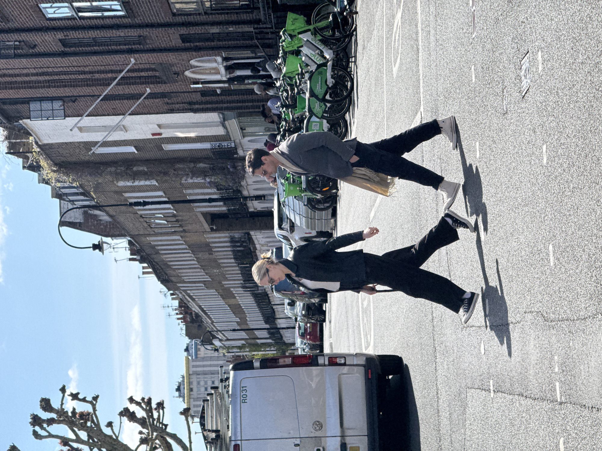 Two individuals are standing on a street in London, United Kingdom. The image is oriented with the street running diagonally from the lower right to the upper left. A silver van is parked in the lower center of the frame. A woman is standing slightly closer to the camera, wearing a dark suit with a white shirt. A man is positioned further away, wearing a grey suit and a light-colored scarf. Behind them, rows of green Lime bikes are parked along the side of the street, adjacent to the building. The buildings are brick and white. The sky is blue with a few clouds. The street has lane markings and some road markings. The lighting indicates it is daytime.