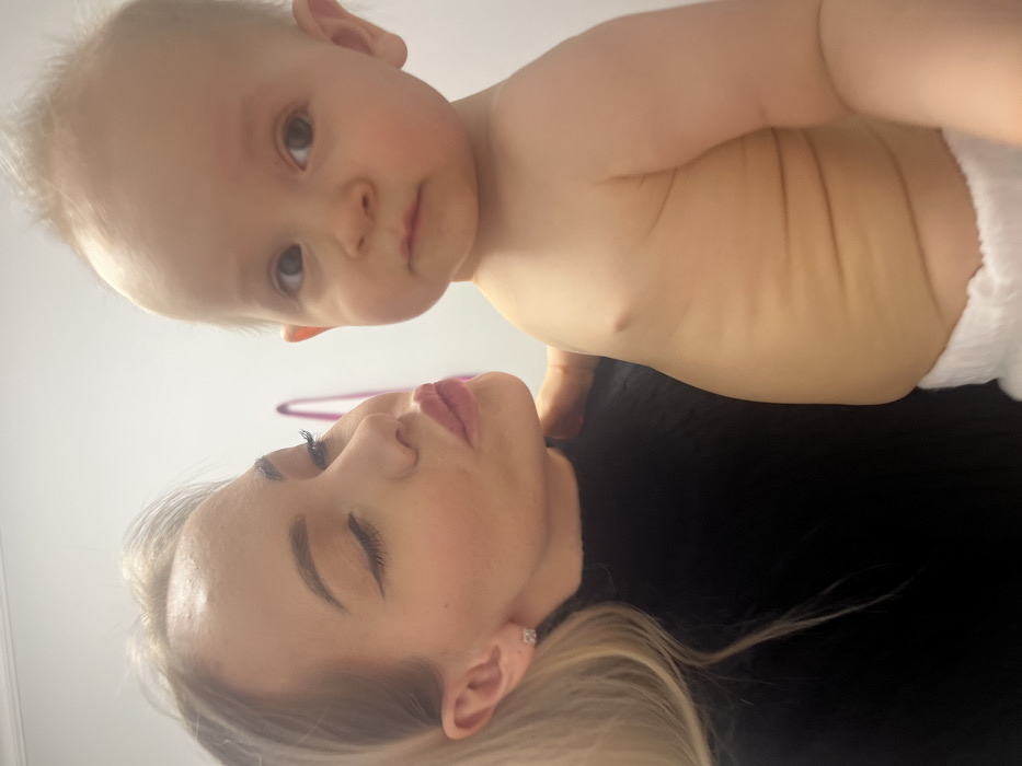 A fair-skinned woman with closed eyes and blonde hair is lying down. Above her is a baby wearing a diaper, peering down at her. The baby has fair skin and light blonde hair. The woman is wearing a black top and has a small earring. The baby's bare chest and stomach are visible, and a hand is seen supporting the baby.