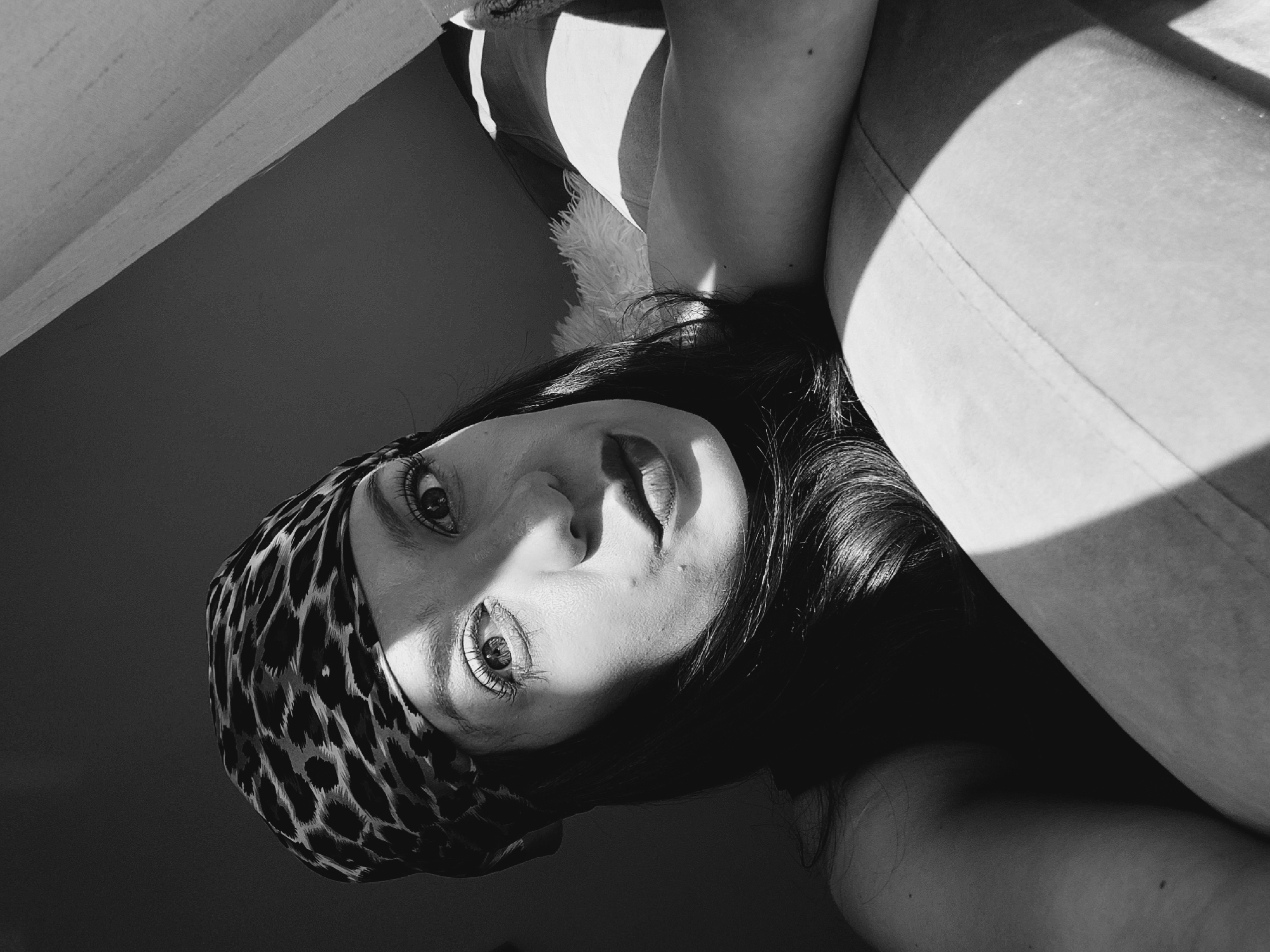 A black and white, close-up, selfie-style photo shows a woman lying down, looking at the camera. She is wearing a leopard print bandana. Her face is partially in shadow from a harsh light source coming from the right. She has dark hair and her lips are painted red. The background is dark. The photo was taken in Sofia, Bulgaria.