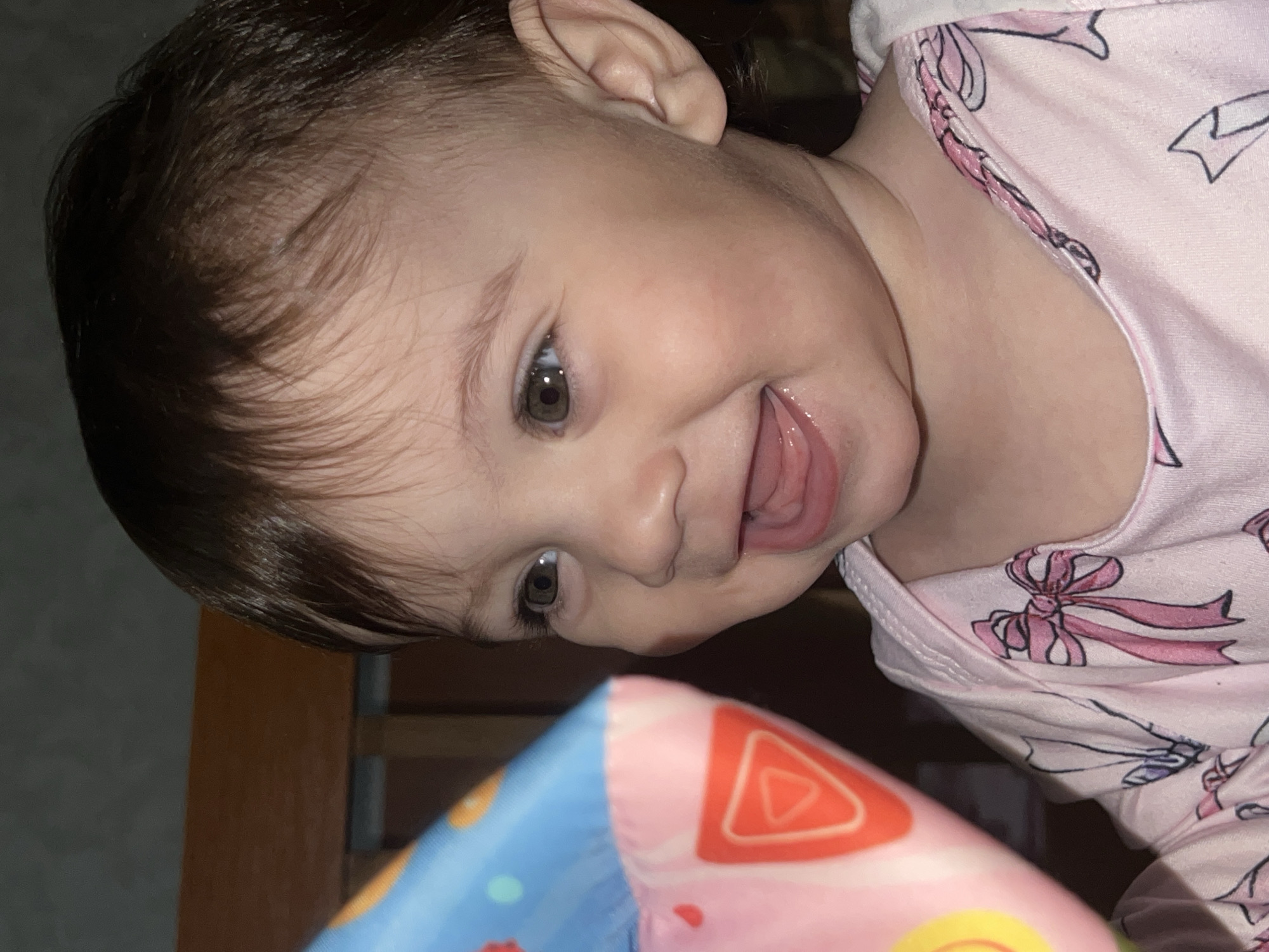 A close-up shot shows a baby with light skin and dark brown hair, wearing a pink shirt with bow patterns. The baby is looking down and to the left with a partially open mouth revealing the tongue. A colorful toy with a red triangle design is partially visible in the lower center of the frame. The background includes a wooden structure. The image does not depict Elena, Bulgaria, or any discernible city location.