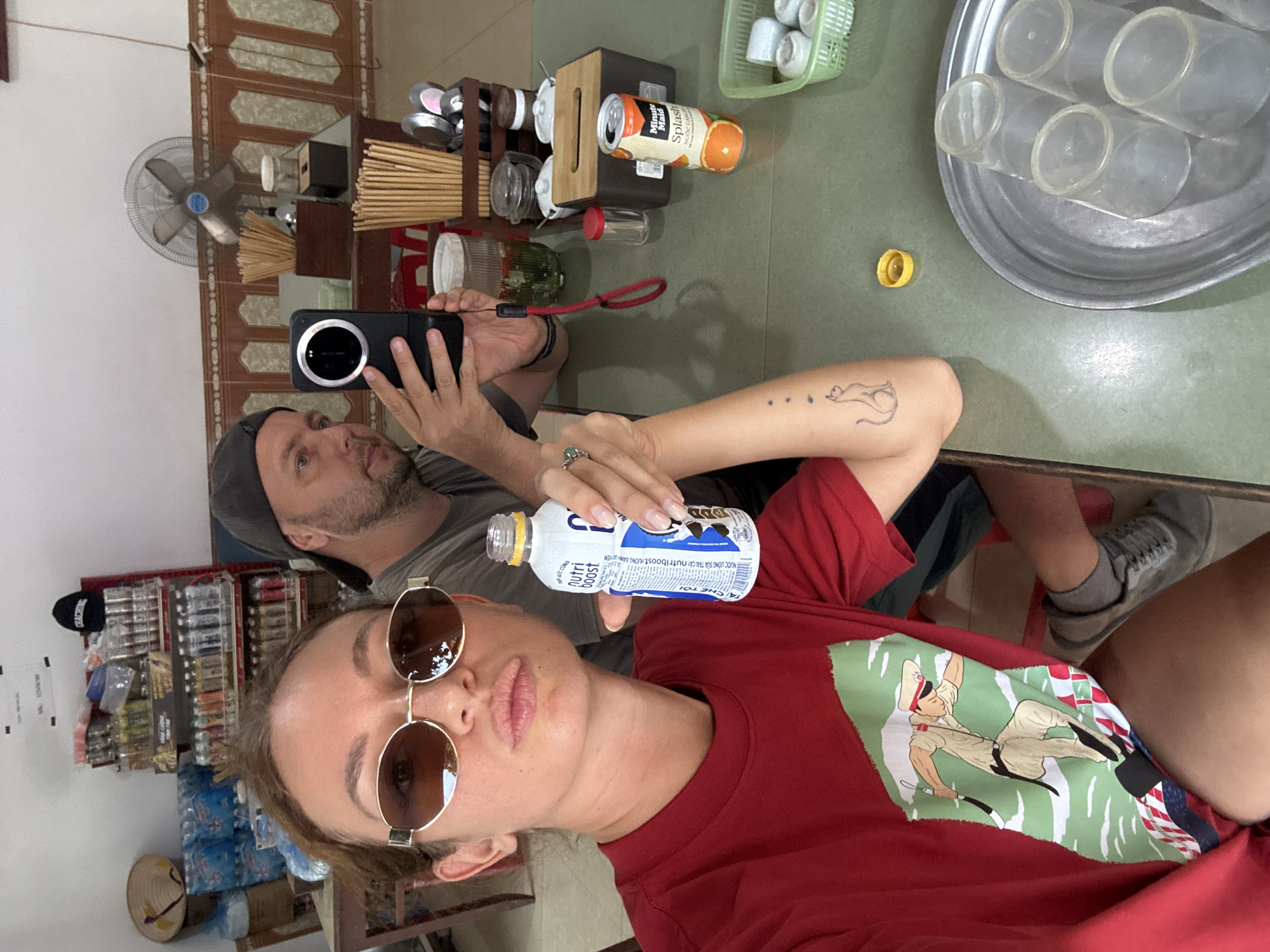 This image, rotated 90 degrees clockwise, captures two individuals inside a casual establishment, likely an eatery or cafe, in Phố Ràng, Vietnam. In the foreground, a woman with sunglasses and a red t-shirt featuring a graphic of a man in uniform is taking a selfie, holding an open bottle of "Nutriboost" drink. She has a distinctive cat tattoo on her right arm. Behind her, a man with a beard and a dark cap is holding a black smartphone with a prominent camera module, looking intently at its screen, possibly filming or viewing content.
The scene is set around a light green tabletop laden with various items. These include a wooden condiment caddy filled with chopsticks, a yellow-capped "Minute Maid Splash" orange juice can, and a metal tray holding several clear plastic cups. In the background, a ceiling fan is mounted on a patterned wall. To the left, shelves are stocked with bottled drinks and other merchandise, and a traditional Vietnamese conical hat (nón lá) is visible.
The visible text on the drink bottle and can includes "SỨC CŨNG", "nutri boost", "DANH KEM", "NƯỚC UỐNG SỮA TRÁI CÂY Nutriboost HUONG B", "Minute Maid Splash", and "NƯỚC CAM". The ambiance suggests a relaxed moment during the day.