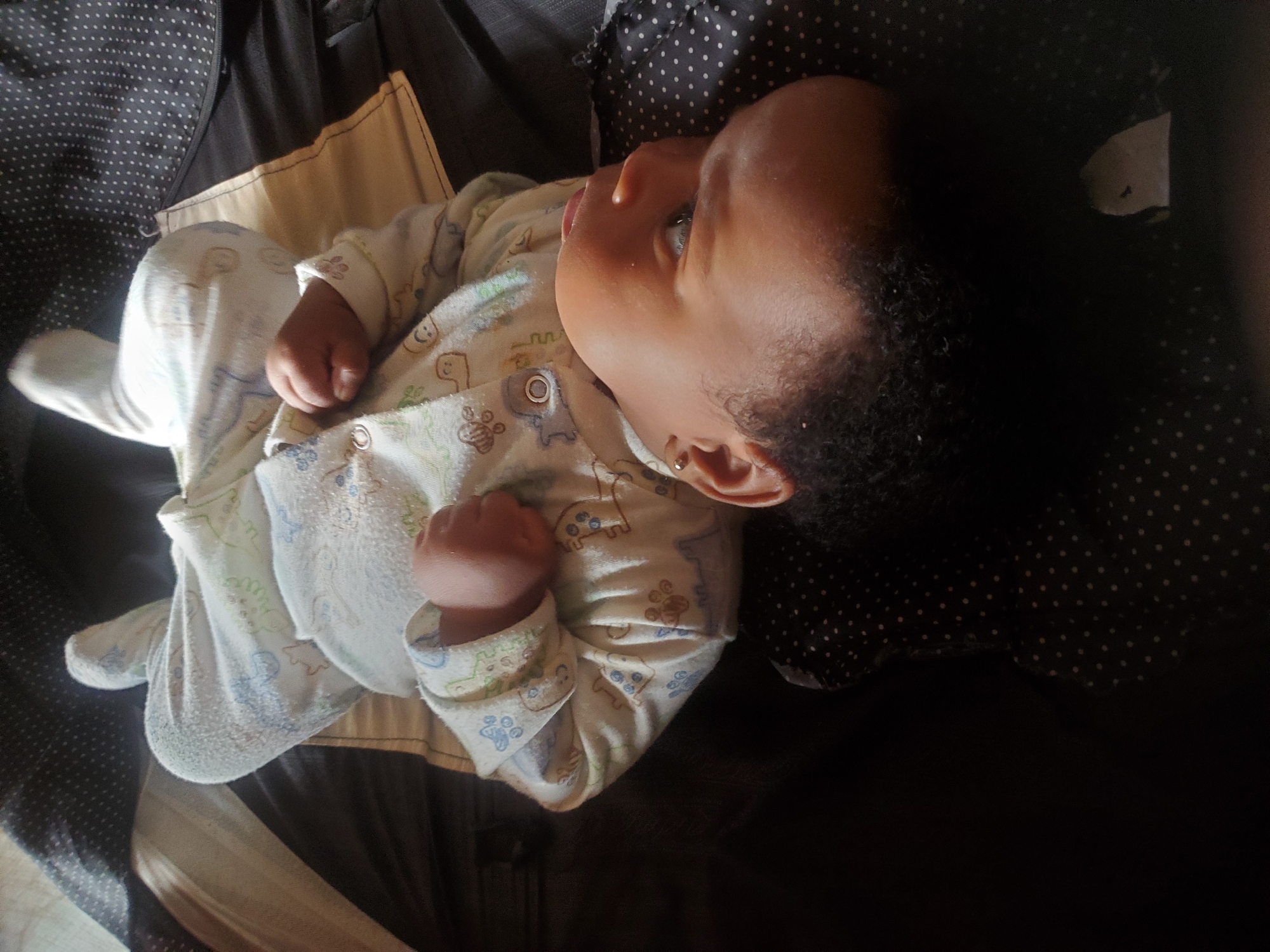 The image shows a baby lying on its back. The baby is wearing a light-colored onesie with a pattern of animals. The baby has dark hair and skin, and appears to be wearing a small earring. The baby is lying on a dark surface with a polka dot patterned object. The lighting appears to be coming from the left, casting shadows on the right side of the image. The image does not have any clear indications about the city of Ukwu-Abwa, Nigeria. The image does not provide forensic information about the location.