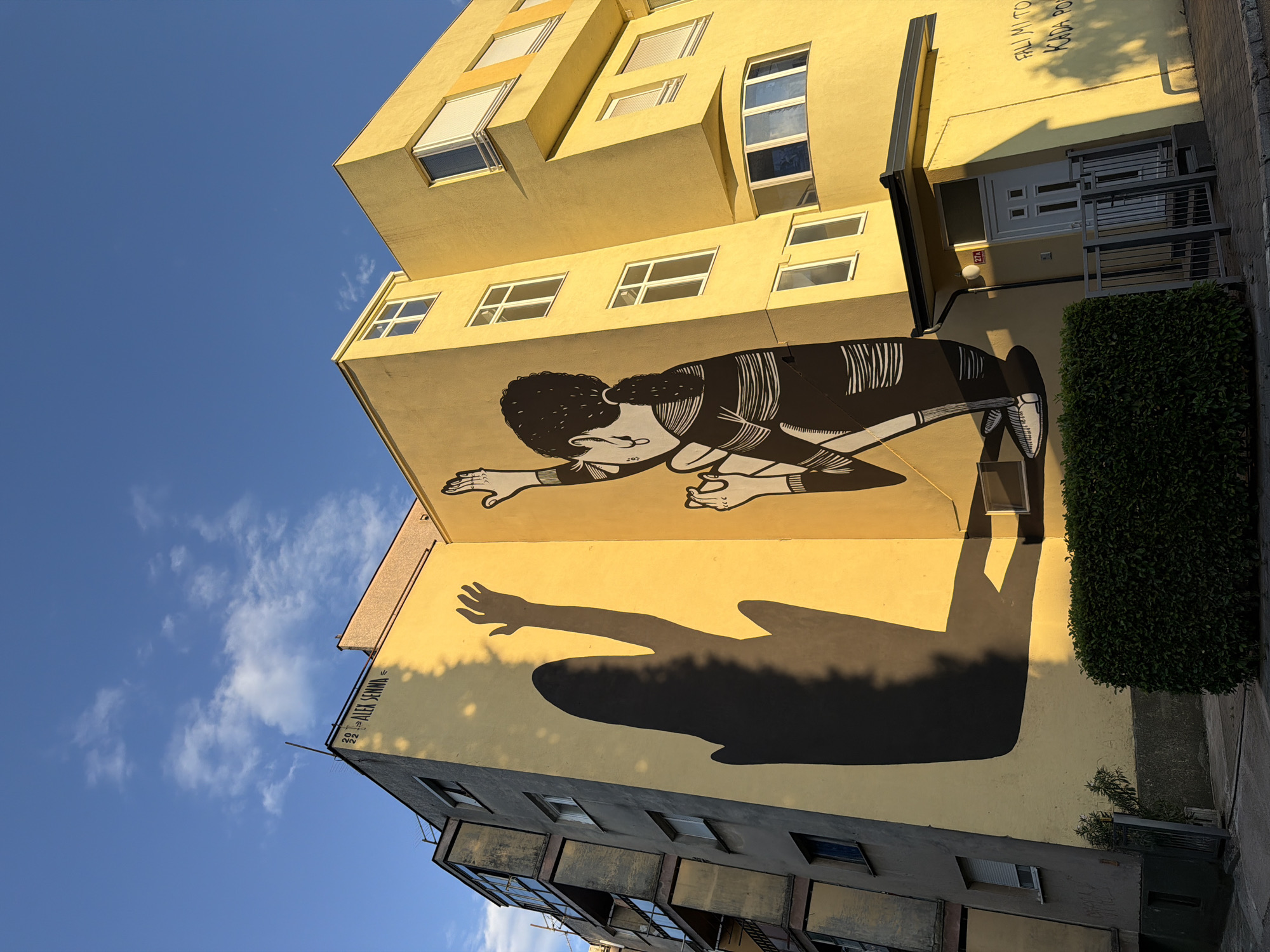 The image depicts a mural on a yellow building in Mostar, Bosnia and Herzegovina. The mural features a black and white illustration of a person with their arms raised and a large shadow cast on the wall. The building is multi-story with visible windows. A portion of an adjacent building, with a more muted color palette, is also visible. The scene is illuminated by sunlight, creating strong shadows. In the foreground, there is a green hedge and a sidewalk with a metal fence and a doorway. The sky above is blue with scattered white clouds. Text is visible on the building, including "2022 ALEK SENNA" and some graffiti in the lower right corner.