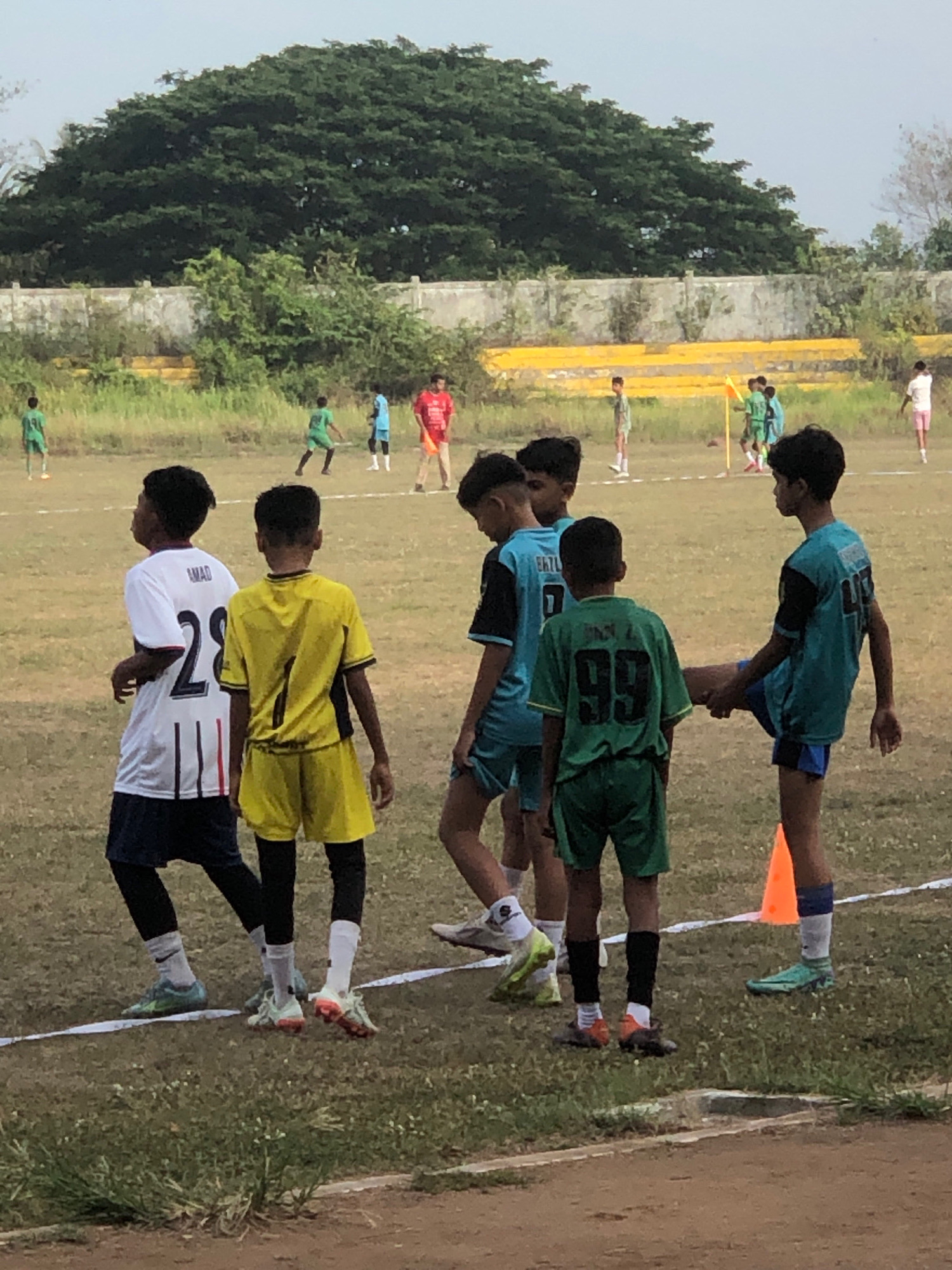 The image depicts a group of young boys in athletic attire on a grassy field, possibly a sports ground. The boys are engaged in what appears to be a casual practice or game of soccer.
In the foreground, five boys are visible. From left to right, the first boy wears a white t-shirt with black and red stripes on the sleeves and the number "28" on the back. He is also wearing dark shorts and black leggings. The second boy is in a yellow jersey with the number "1" on the back, yellow shorts, and black leggings. The third boy, partially obscured, wears a light blue jersey. The fourth boy is dressed in a green jersey with the number "99" on the back and green shorts. The fifth boy wears a teal jersey with the number "47" on the back and blue shorts, and he is shown with his right leg extended, seemingly interacting with another player or stretching.
In the background, several other individuals are present on the field, including some wearing red, green, and light blue attire, suggesting they are also participants in some form of sporting activity. There are also yellow benches or bleachers visible in the distance, along with a large, dense tree canopy and a concrete wall. The ground is a mix of dry grass and dirt.
The overall atmosphere suggests an outdoor sports event or training session. The image was taken in Lhokseumawe, Indonesia.