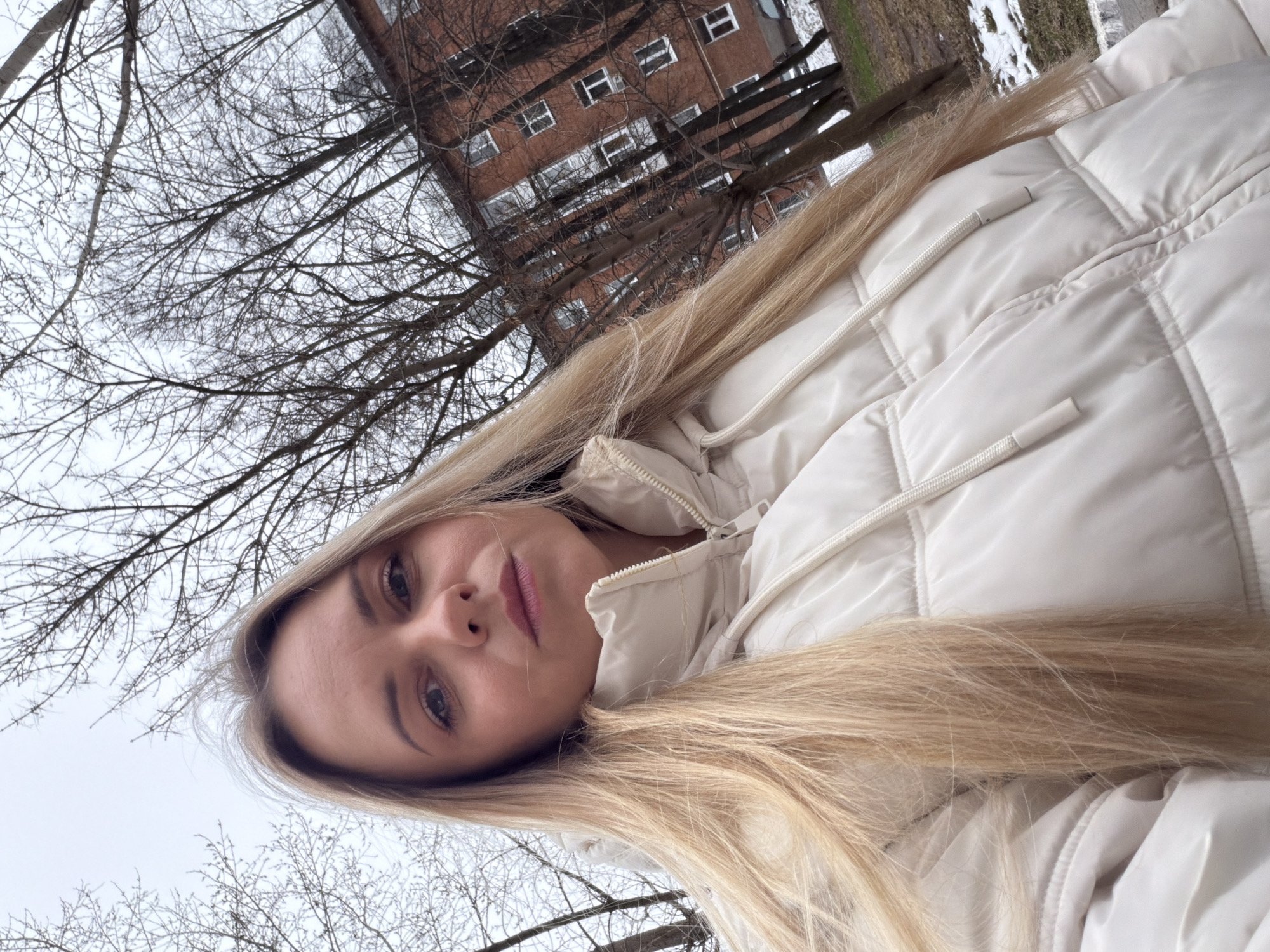 A blonde woman wearing a white puffer jacket is posing for a selfie outdoors. Bare trees are visible in the background, as is a multi-story brick building. Based on the architecture and sparse vegetation, the photo may have been taken in a park in late fall or early winter. There is a patch of snow on the ground. Without additional visual information, pinpointing a specific location in Votkinsk, Russia, is not possible.