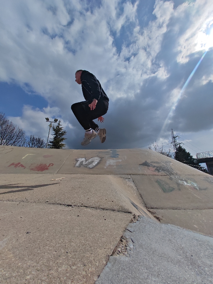 The image captures an outdoor urban concrete environment in Shumen, Bulgaria, under a partly cloudy sky with visible sunlight and lens flare originating from the upper right. A single male individual is depicted mid-air in a crouched jumping posture, facing left. He wears a black tracksuit with white stripe detailing on the sleeves and pant cuffs, along with light-colored sneakers and white socks. His hands are drawn close to his torso, and his mouth is slightly open. The subject is positioned above a concrete structure consistent with a skatepark, featuring angled surfaces, visible cracks, and multi-colored graffiti including red characters (e.g., "НАРО") and a large white "M". A small, light-colored stick rests on the flat concrete surface in the foreground. The background includes deciduous trees on the far left, a dual-head streetlight on a pole, a conifer tree, a metal power pylon, and a building or structure with a white railing on the far right. The scene is illuminated by natural daylight.