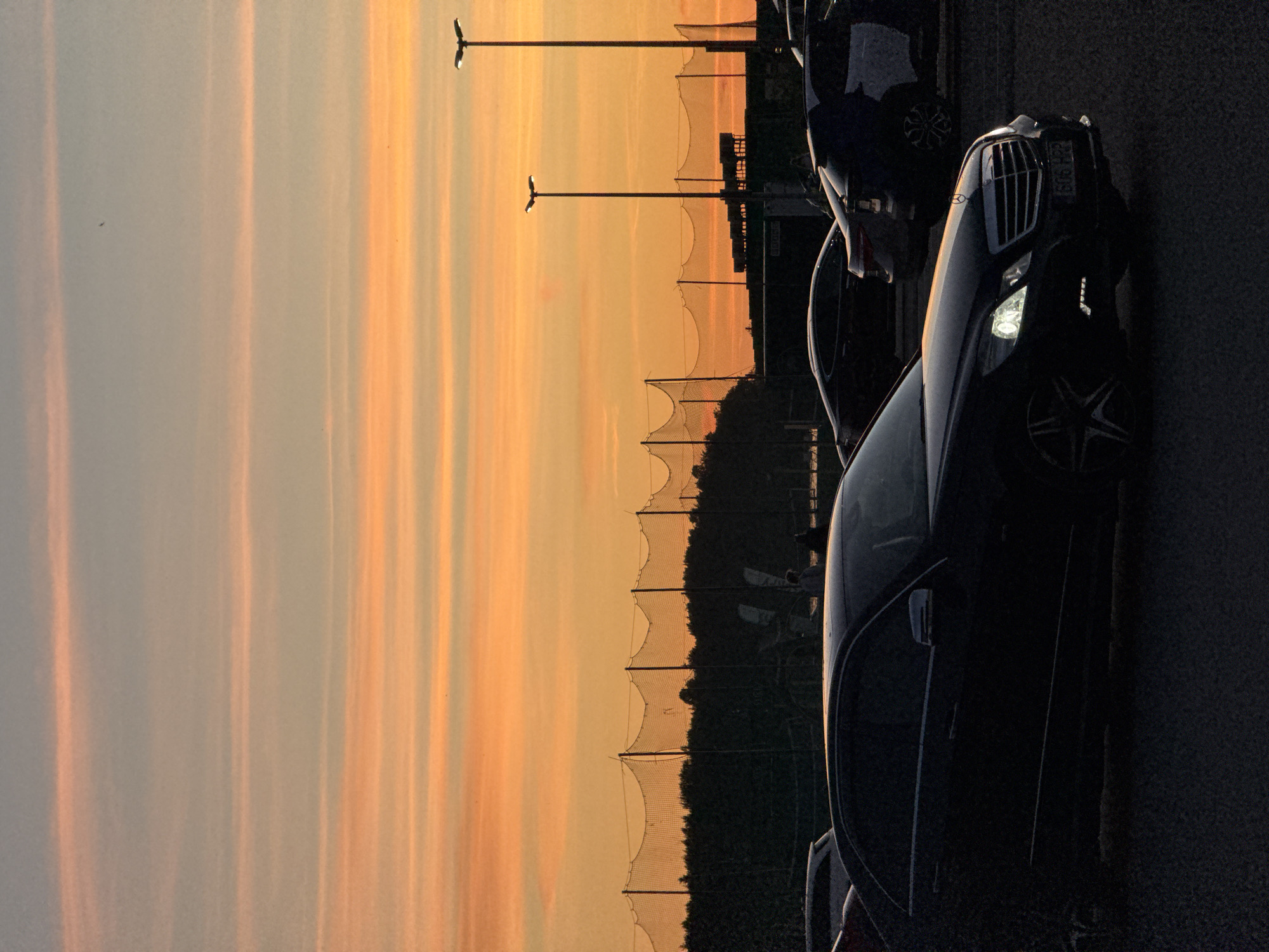 The image captures a sunset over a parking lot in Tallinn, Estonia. In the foreground, a black Mercedes-Benz E-Class sedan is prominently featured, its headlights illuminated. Several other vehicles are parked in the lot. In the background, a sports net encloses an area, likely a driving range or a similar facility, with a dense line of trees behind it. Two tall lampposts stand against the colorful sky, which is streaked with orange and pink hues from the setting sun. A few distant figures are visible, silhouetted against the fading light.