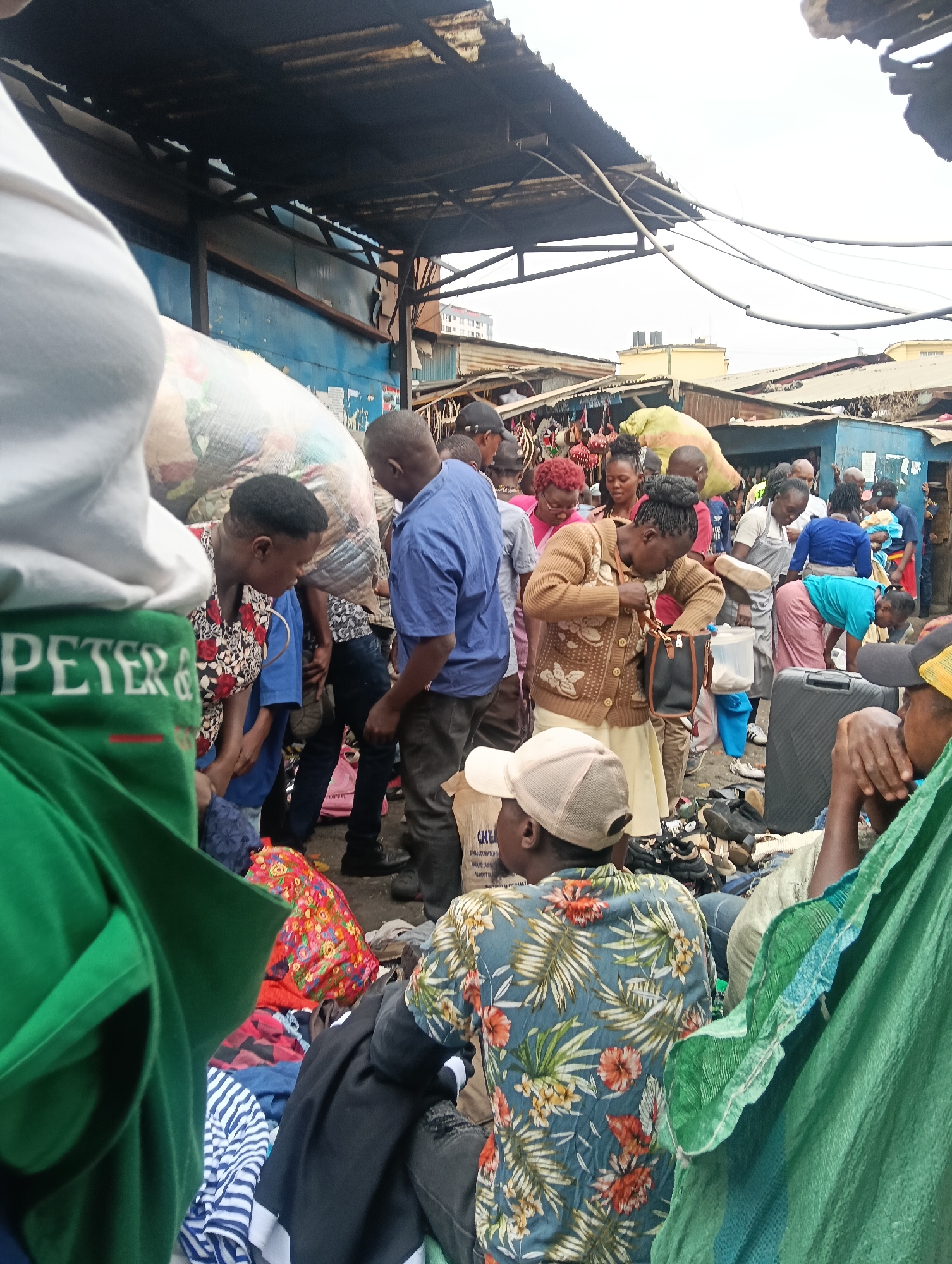 The image depicts a busy outdoor market scene in Nairobi, Kenya, likely a flea market or a street market. A dense crowd of people are engaged in shopping and interacting. The environment is characterized by makeshift stalls and shelters with corrugated iron roofing, suggesting an informal commercial setting. Clothing, bags, and various merchandise are visible, indicating a variety of goods being sold. The people in the image are of diverse ages and ethnicities, dressed in casual clothing typical of a warm climate. Some individuals are examining goods, while others appear to be conversing. The overall atmosphere suggests a vibrant and bustling marketplace.