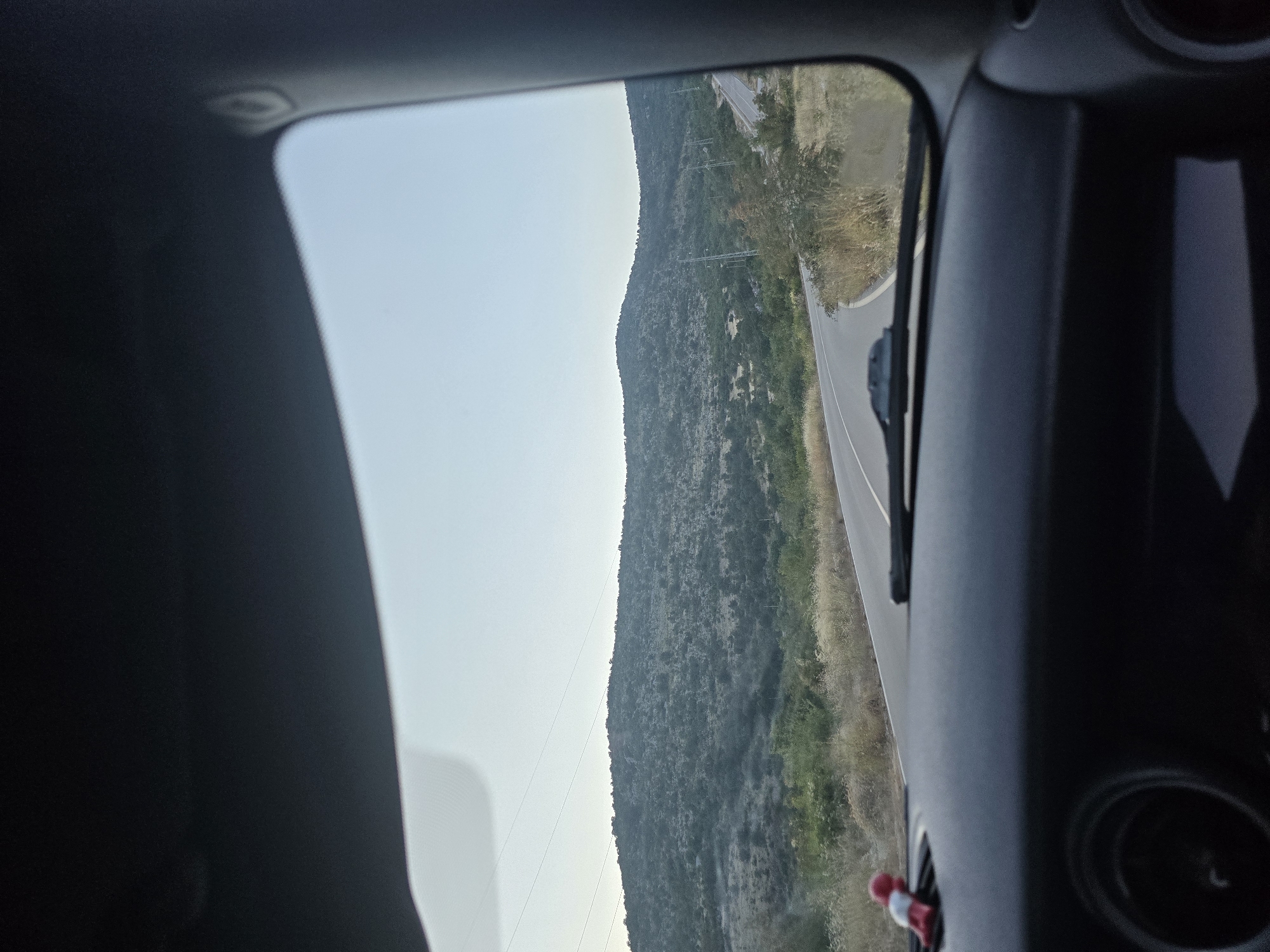 The image is a view from inside a car driving on a paved road in a mountainous, rural area. The road curves to the right, leading towards a large, scrub-covered hill. The sky is clear and pale blue, suggesting it might be late afternoon or early morning. Power lines are visible stretching across the horizon. Inside the car, the dashboard is visible, with a small red and white figurine attached to the left side. The windshield wipers are also visible on the windshield. The location is specified as the city of Klopot, Montenegro.