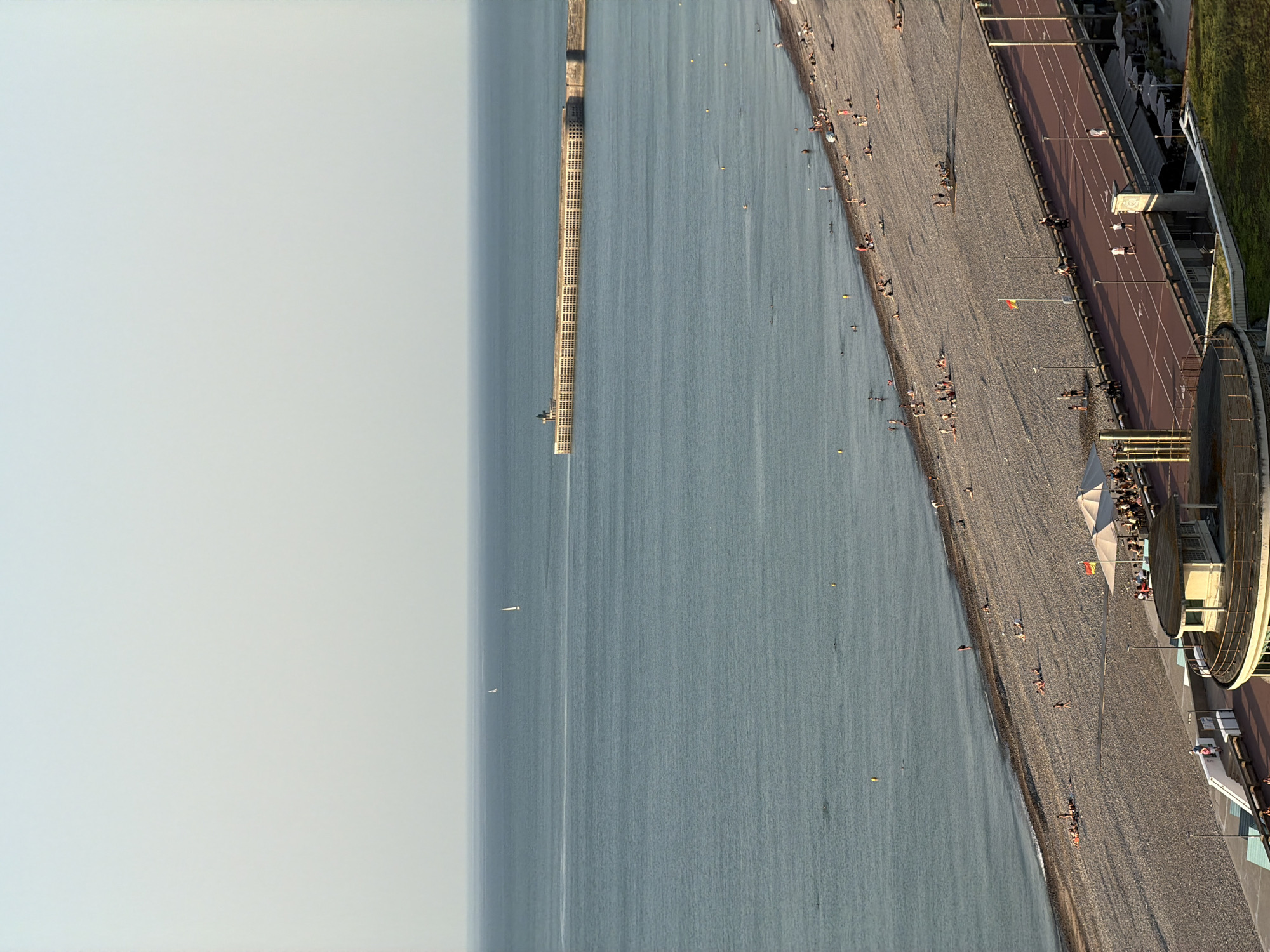 This is an aerial, full-shot, vertical, landscape image taken from a high vantage point, looking down at a beach and the sea. The scene depicts a sunny day at a pebble beach lined with a promenade, where people are engaged in various activities.
In the water, a long, rectangular structure, likely a pier or breakwater, extends from the shore into the sea. There are a few small boats visible in the water, and two even smaller boats or buoys further out. The sea itself is a calm, blue-grey color.
The beach is covered in grey pebbles, and scattered across it are numerous people, some sunbathing, others walking. A paved promenade runs parallel to the beach, with a red track on it, suggesting it may be used for running or cycling. More people are seen on the promenade, and there are lamp posts and a few small structures, including what appears to be a building with a rounded section. Some beach umbrellas are set up, and a flag is visible on a flagpole. The overall atmosphere suggests a leisurely summer day.
The image is well-lit, suggesting it was taken during daylight hours, likely in the late afternoon or early evening given the warm tones on the promenade and the long shadows cast by some of the structures and people. The sky is a light, hazy blue, indicating clear weather. The location is Dieppe, France, as indicated by the context. No visible text is present in the image.