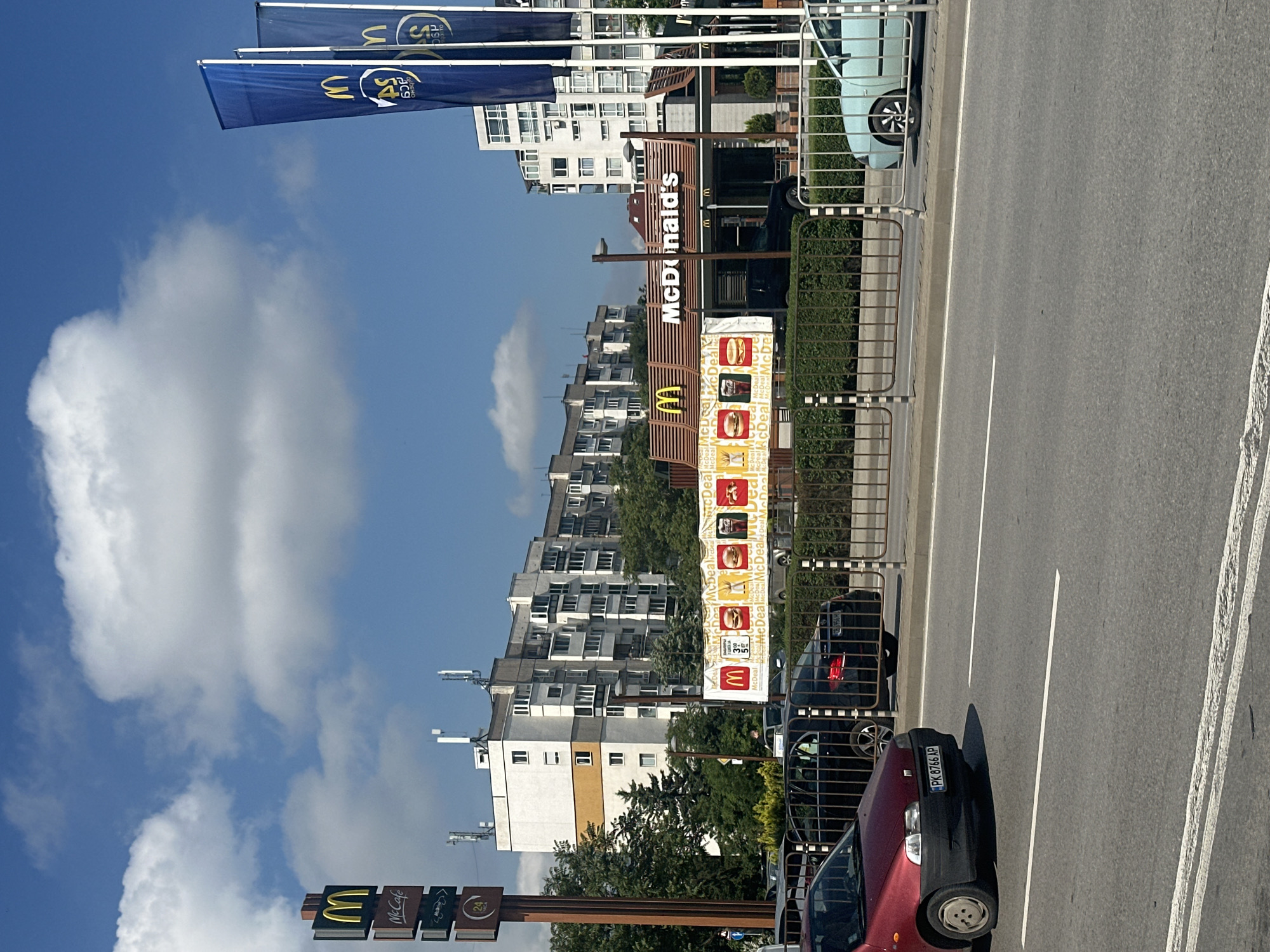This urban scene depicts a McDonald's restaurant and its surroundings under a bright, sunny sky with scattered white clouds. The image captures a vibrant daytime environment, likely late morning or afternoon, indicated by the strong sunlight and shadows.
Prominently featured on the left side is a McDonald's location. A large vertical sign clearly displays "McDonald's" in white lettering on a brown background. Adjacent to it, a tall pole sign shows the golden "M" logo, "McCafé," and a "24" symbol, suggesting 24-hour service. A drive-thru menu board is visible, advertising various "McDeal" promotions with images of burgers, fries, and drinks, repeating the text "McDeal." Overhead, two blue flags with the McDonald's "M" logo and "40" (likely indicating "40 years" or an anniversary) are flying.
A multi-lane road occupies the right side of the image, separated from the restaurant area by a metal safety barrier. Several vehicles are visible; in the foreground, a dark red hatchback car with the Bulgarian license plate "PK 8766 AP" is partially seen next to the road. Further back, a light blue compact car is parked near the McDonald's.
The background is dominated by several multi-story residential apartment buildings, some featuring balconies and one with a distinctive yellow and white facade. Rooftop antennas are visible on some of these structures. Green trees and bushes are interspersed throughout the setting. The presence of the Bulgarian license plate "PK 8766 AP" confirms the location in Bulgaria, specifically matching the context of Sofia.
Visible text includes: "McDonald's," "Mc Cafe," "McDeal" (repeatedly), "PK 8766 AP," the "M" logo, and "40" on the flags.