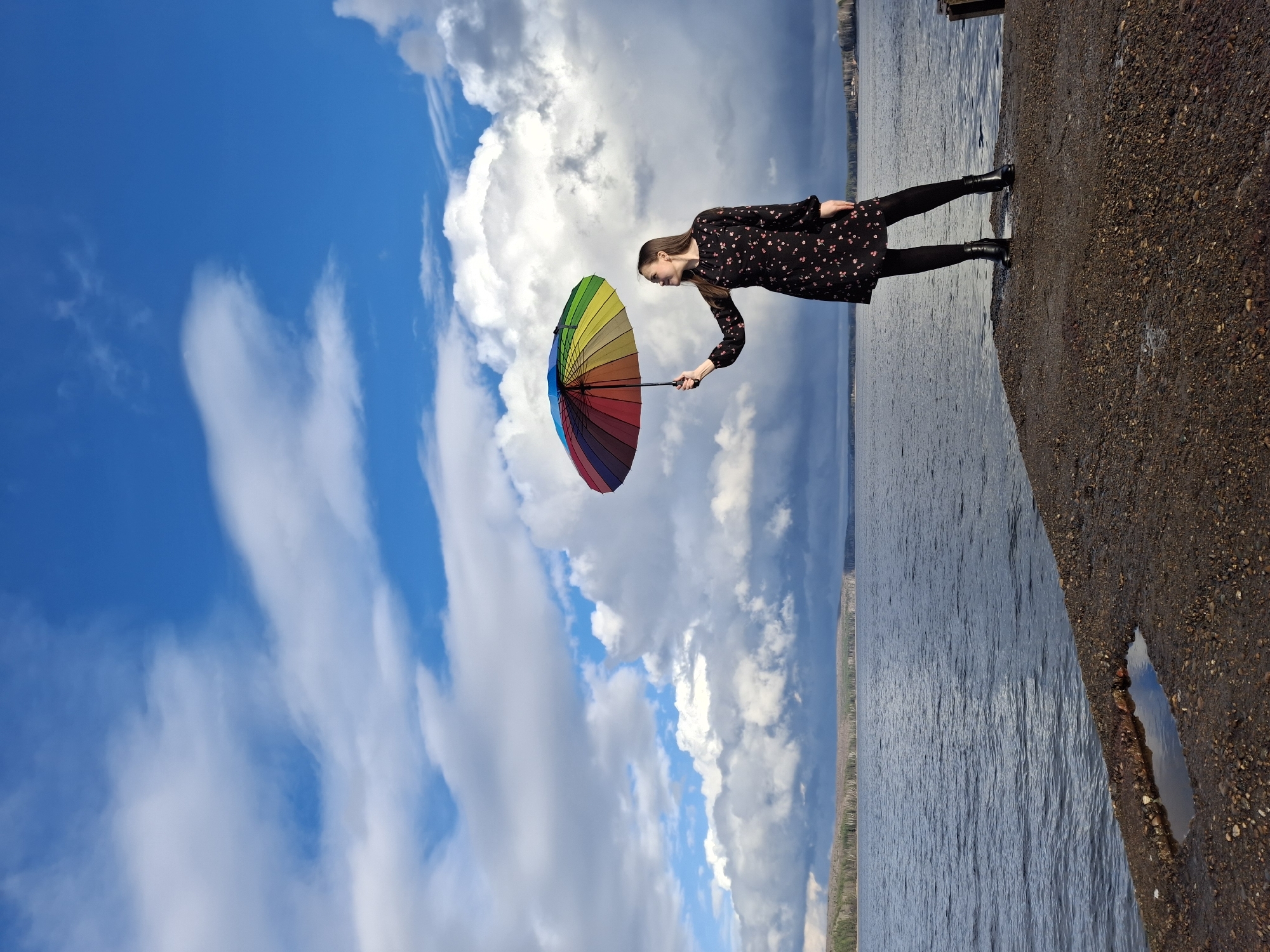 A vertical medium shot depicts a woman holding a rainbow-colored umbrella against a backdrop of a blue and cloudy sky and a water body. The woman is wearing a dark dress with small floral patterns, black tights, and black boots. She is standing on a paved area, likely a sidewalk or embankment. The right side of the image shows water and a shoreline, indicating a riverside location in the city of Chaikovsky, Russia.