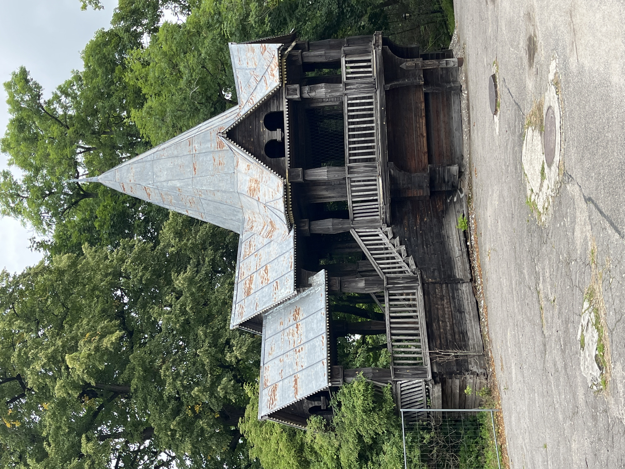 An aged, ornate wooden structure, possibly a gazebo or pavilion, stands in a park-like setting in Kaliningrad, Russia. The structure features a multi-tiered, conical metal roof with visible signs of rust and age. Elaborate wooden carvings adorn the eaves and pillars. A wooden staircase with railings leads up to the main platform, which is also enclosed by a wooden railing. The building is surrounded by lush green trees and foliage. The ground in front of the structure is paved with cracked asphalt, showing signs of wear and tear, with a couple of manhole covers visible. A metal fence encloses a section of the area to the left of the structure, suggesting ongoing maintenance or restricted access. The overall impression is one of historical charm, albeit with evident signs of weathering and the passage of time.
