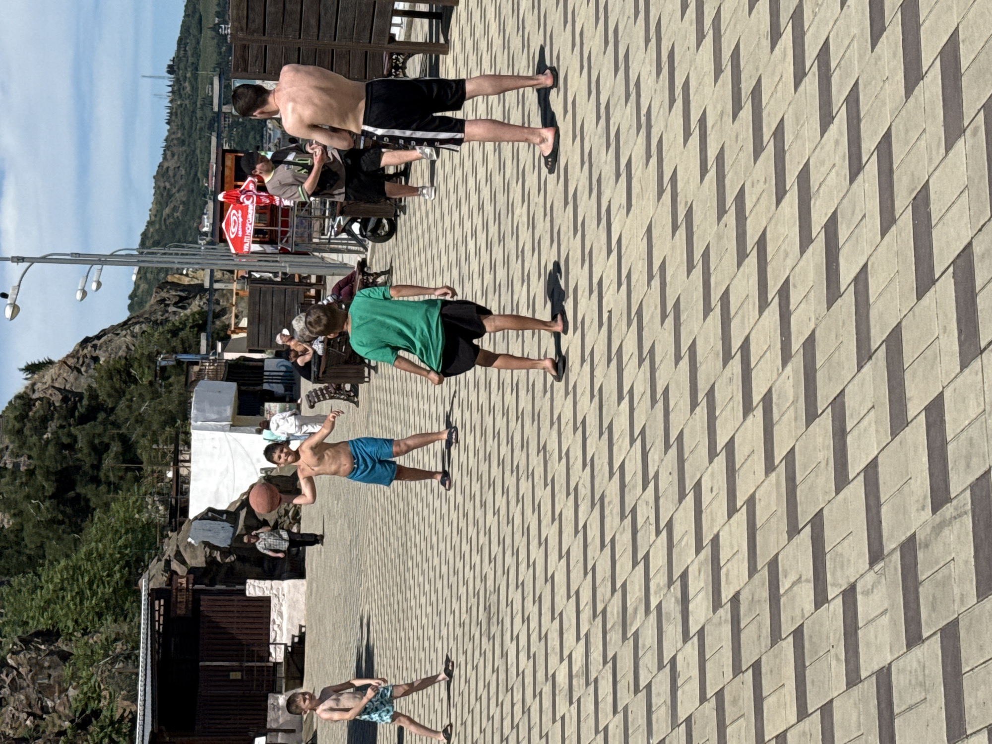 The image, rotated 90 degrees counter-clockwise, depicts a sunny outdoor promenade alongside rocky, tree-covered hills under a clear blue sky.
In the foreground, two young males are visible on the light-colored, geometrically patterned paved surface. One, shirtless and wearing blue shorts, holds a basketball under his left arm. The other, standing to the right, wears patterned swim trunks. Further back, a shirtless young man in black shorts and flip-flops walks away from the viewer, casting a long shadow. Another male, wearing a green t-shirt and black shorts, also walks away, casting a similar shadow.
To the left of the promenade, a rocky hillside covered in trees and scrub brush rises, with various buildings and structures nestled into the landscape. In the midground, along the right side of the path, several people are gathered around tables, some seated under a large red umbrella. The umbrella features visible Cyrillic script, part of which appears to be "ХОЛОДНОЕ" (Kholodnoe), suggesting an advertisement for cold items.
The strong, distinct shadows cast by the people and structures indicate a sunny day, likely mid-day or afternoon. The attire of the individuals suggests a warm, leisure-focused environment, possibly a resort area or beach promenade. The overall scene conveys a relaxed, summery atmosphere.