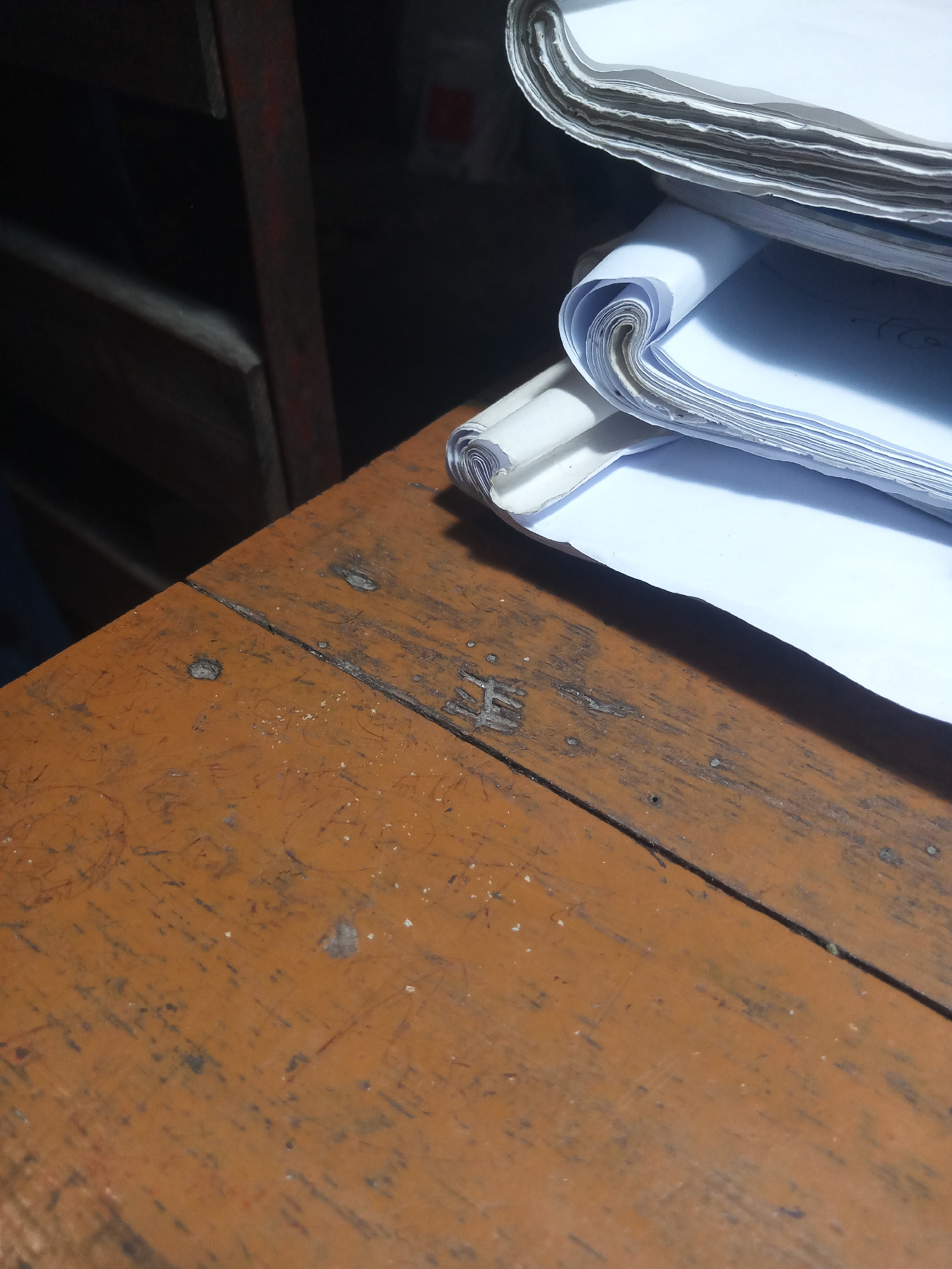 The image shows a close-up of a worn, orange-brown wooden surface, possibly a desk or table. There are faint scratches and marks on the surface, suggesting age and use. A faint imprint of what appears to be a character or symbol is visible on the wood. To the right, a stack of white papers or documents is piled high, with some edges curling and showing multiple layers. In the background, a dark, out-of-focus area suggests a shelf or storage unit with red and dark elements. The lighting is uneven, casting shadows and highlighting certain areas. The image was taken in Hathazari Upazila, Bangladesh.