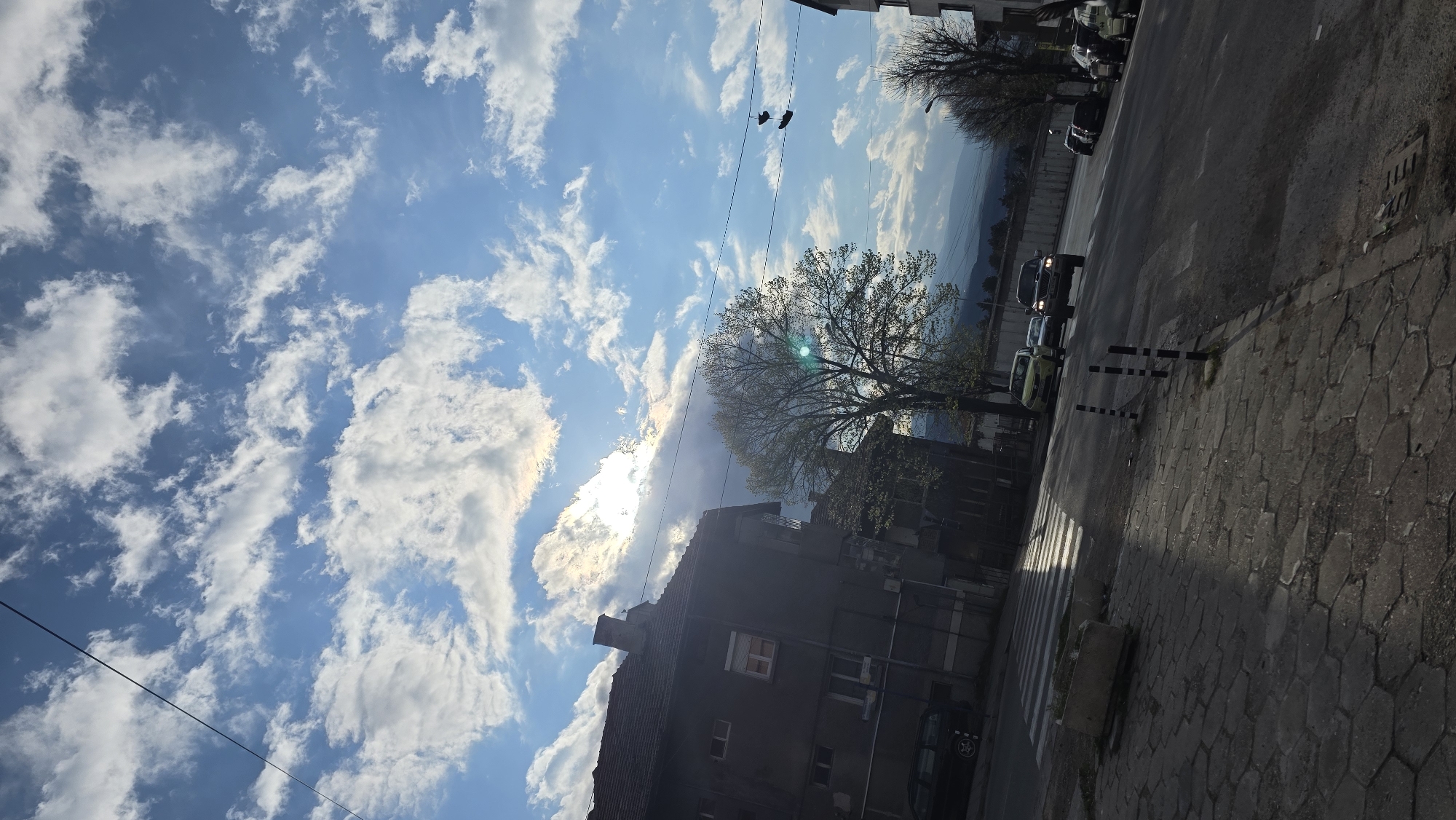 The image captures a street scene in Blagoevgrad, Bulgaria. The view is from the sidewalk, looking towards a street with several parked and moving vehicles. The street runs adjacent to a building, likely residential or commercial, with a mature tree in front partially obscuring it. Overhead, a bright, partly cloudy sky dominates the scene. A pair of shoes hang from a power line. In the background are mountains in the distance. The perspective of the photo is slightly tilted counterclockwise.