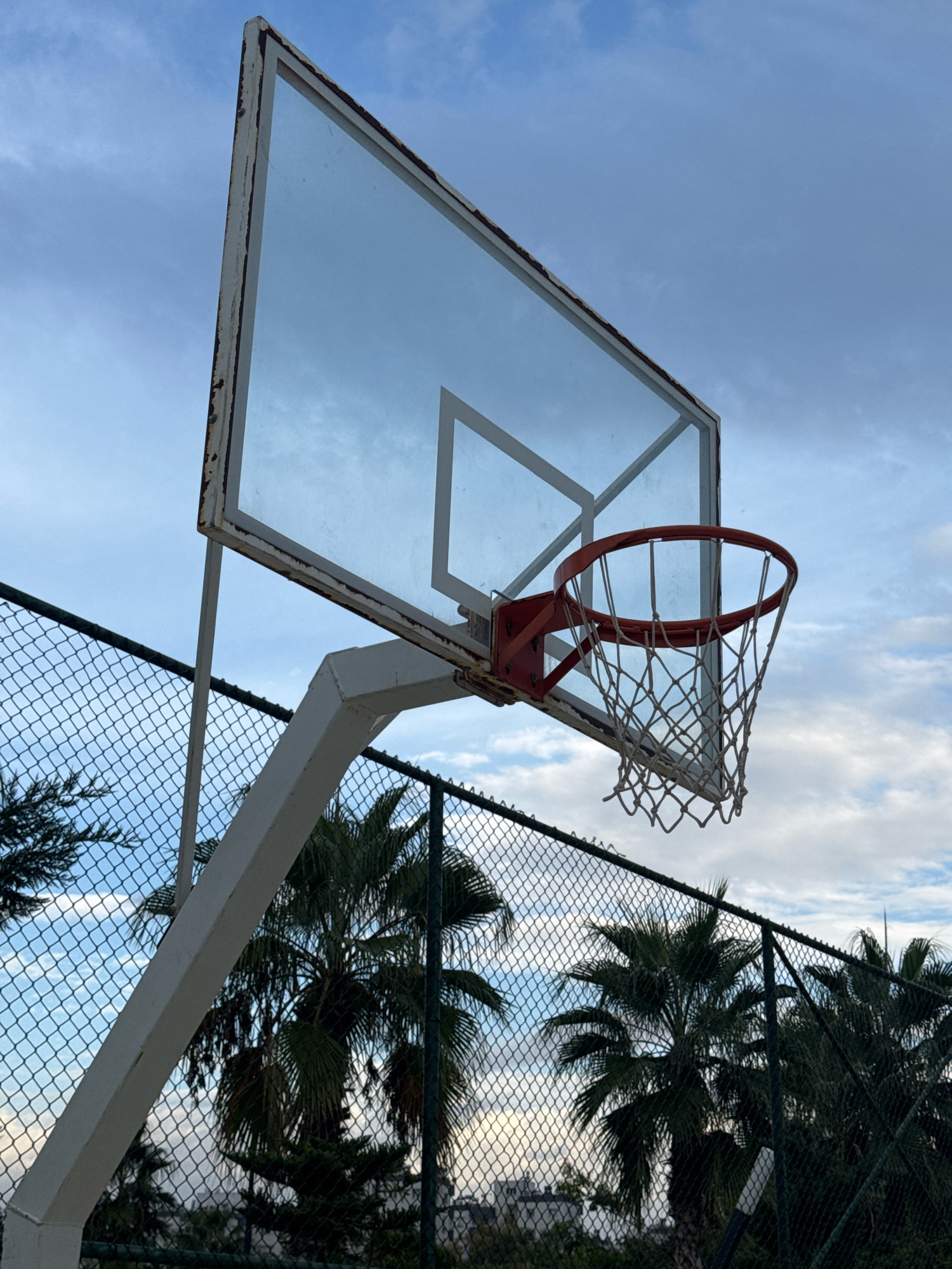 This image shows a basketball hoop and backboard against a cloudy sky. The basketball hoop is red and has a white net. The backboard is made of glass and has a white frame that shows signs of rust. The hoop is attached to a white metal pole. In the background, there is a chain-link fence and palm trees, suggesting a warm climate. The scene is likely outdoors in a park or recreational area. The description does not specify a precise location within Antalya, Turkey, but implies a general outdoor setting typical of the city. There are no people or actions depicted in the image.