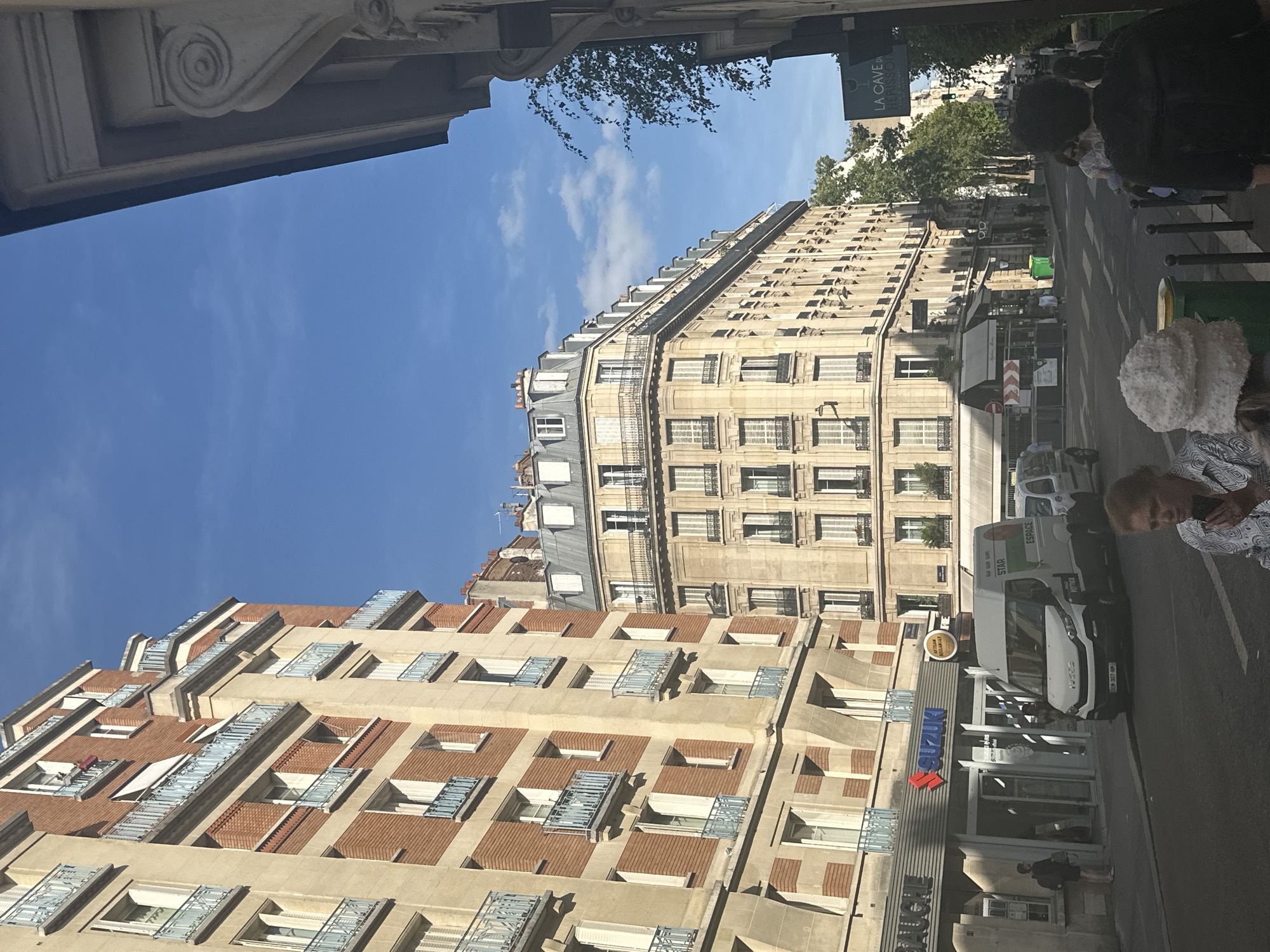 The image depicts a street scene in Paris, France, likely in an urban residential and commercial area. The perspective is from a street level, looking up at several multi-story buildings. The architecture is typical of Parisian buildings, featuring cream-colored stone facades, brickwork, and numerous windows with decorative balconies and shutters.
On the left, a building displays a combination of brick and stone, with repeating light blue balconies. Below this building, there is a storefront with a prominent "SUZUKI" logo. Adjacent to the Suzuki store is another establishment with a sign partially visible that reads "SOUCHE".
In the center of the image, a larger, cream-colored building with a mansard roof and dormer windows dominates the mid-ground. This building has a curved facade as it turns a corner. The street level of this building also appears to have shops or businesses.
A white van with various logos and text is parked on the street in front of one of the stores. Several people are visible on the sidewalk and street. In the foreground, a woman is looking down at her phone, wearing a patterned shirt. Another person wearing a white fluffy hat is partially visible. Further down the street, other pedestrians are walking.
The sky is a bright blue with some scattered white clouds. Trees and some green foliage are visible along the right side of the street, suggesting a leafy environment. A street lamp is visible on the left side of the street in front of the brick building. A dark sign with white text and a graphic of a wine glass hangs on the right side of the street, identifying a location as "LA CAVE DU VERISSON". The overall impression is a sunny day in a bustling Parisian neighborhood.