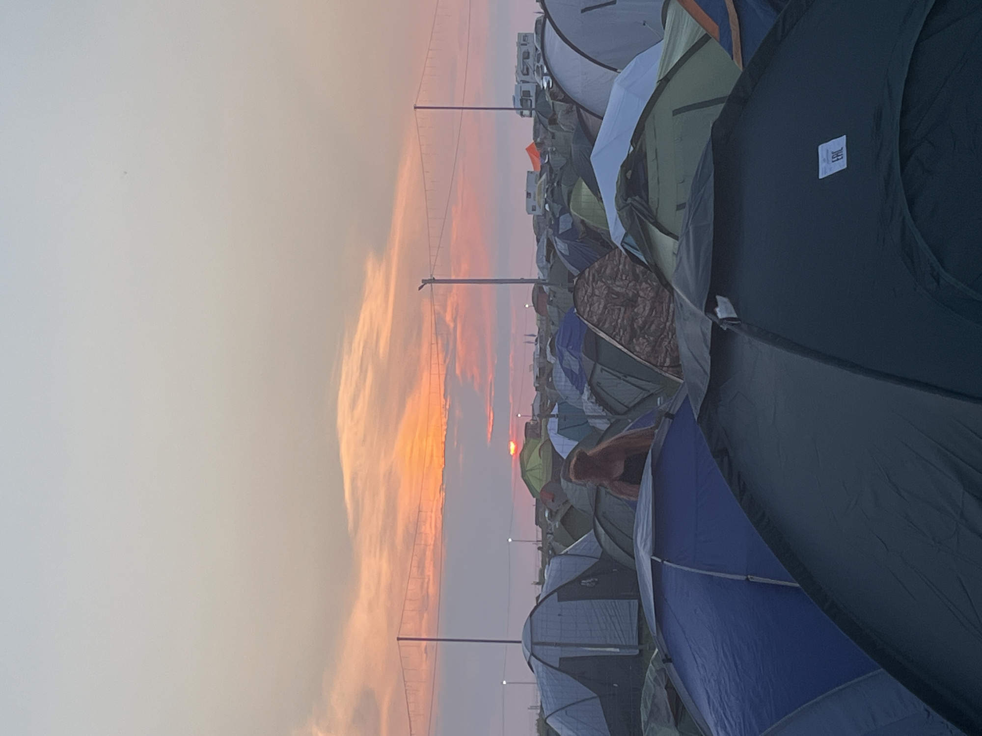 The image depicts a large campsite at sunset, presumably at the Surakovo festival grounds in Russia. The sky is filled with colorful clouds, transitioning from pale blue to orange and pink as the sun sets near the horizon. Numerous tents of various colors and sizes are pitched close together, forming a dense camping area. In the mid-ground, a person with long, reddish hair is standing, looking towards the sunset. In the background, several RVs and trailers are visible, along with light poles and string lights, suggesting a festival or event setting. The overall scene conveys a sense of an outdoor gathering at dusk.