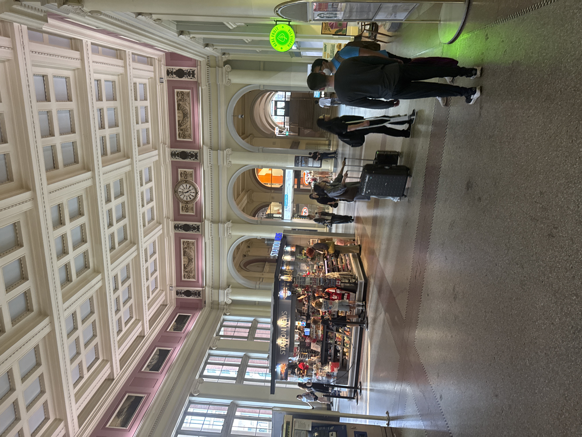 The image depicts the interior of a public transportation station, likely a train or bus terminal, given the presence of a "STATION NEWS" kiosk and people with luggage. The architecture is grand, featuring a high, coffered ceiling with intricate molding and large arched doorways leading to other parts of the building. The walls are adorned with decorative panels and columns. Natural light streams in from tall windows.
In the foreground, several individuals are visible. Two men are standing together, one wearing a dark grey long-sleeved shirt and black pants, the other in a blue shirt and black pants. To their left, a woman with a cast on her leg and holding crutches walks with another person. Further back, a group of people are gathered around the "STATION NEWS" kiosk, which displays a variety of products.
The floor is paved with speckled terrazzo, with inlaid patterns. To the right, a bright green circular sign that reads "Real Fruit Bubble Tea" is prominently displayed, suggesting a food or beverage vendor within the station. The overall atmosphere suggests a busy transit hub. The location is Vancouver, Canada, specifically within Pacific Central Station, evidenced by the distinctive architecture and the "STATION NEWS" outlet common in such transportation hubs.