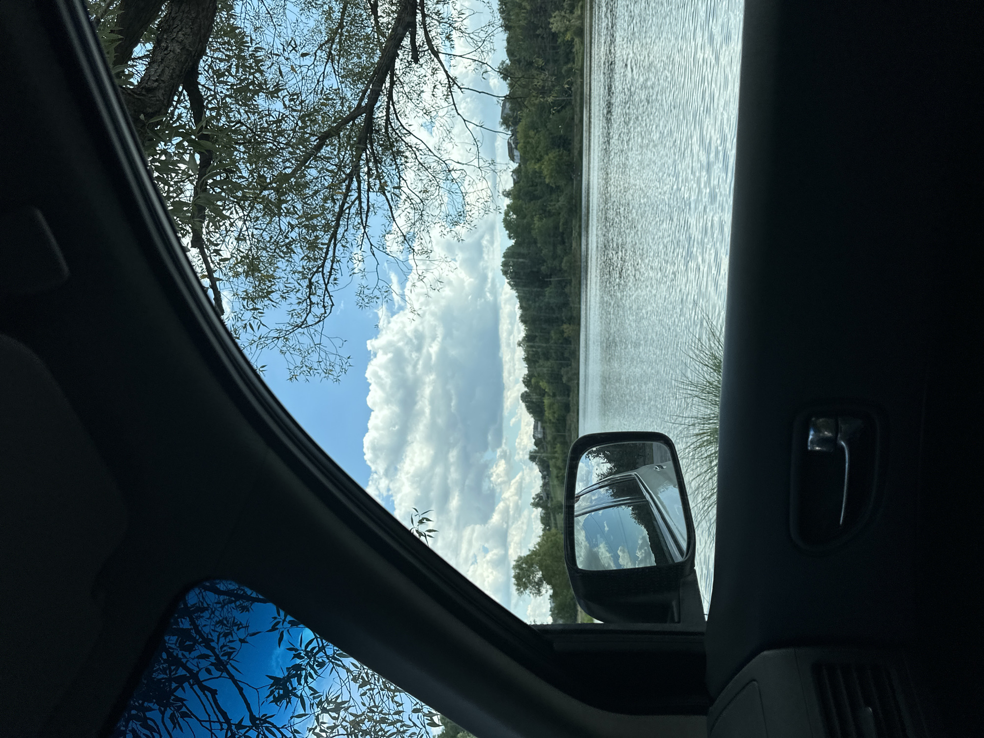 This image captures a serene view of a lake from inside a car, with the window framing the scene. The car's interior, including a door handle and a side mirror reflecting the sky and trees, is visible in the foreground. Outside, the calm lake reflects the bright blue sky dotted with white clouds. A lush green treeline borders the far bank of the lake, with a few houses peeking through the foliage, suggesting a semi-rural or suburban setting. The branches of a large tree hang into the frame from the top right, dappling the scene with shadows. The presence of a car implies this is a scene observed from a vehicle on a road near the lake. The overall atmosphere is peaceful and natural. Given the prompt, this scene is located in Makiv, Ukraine.