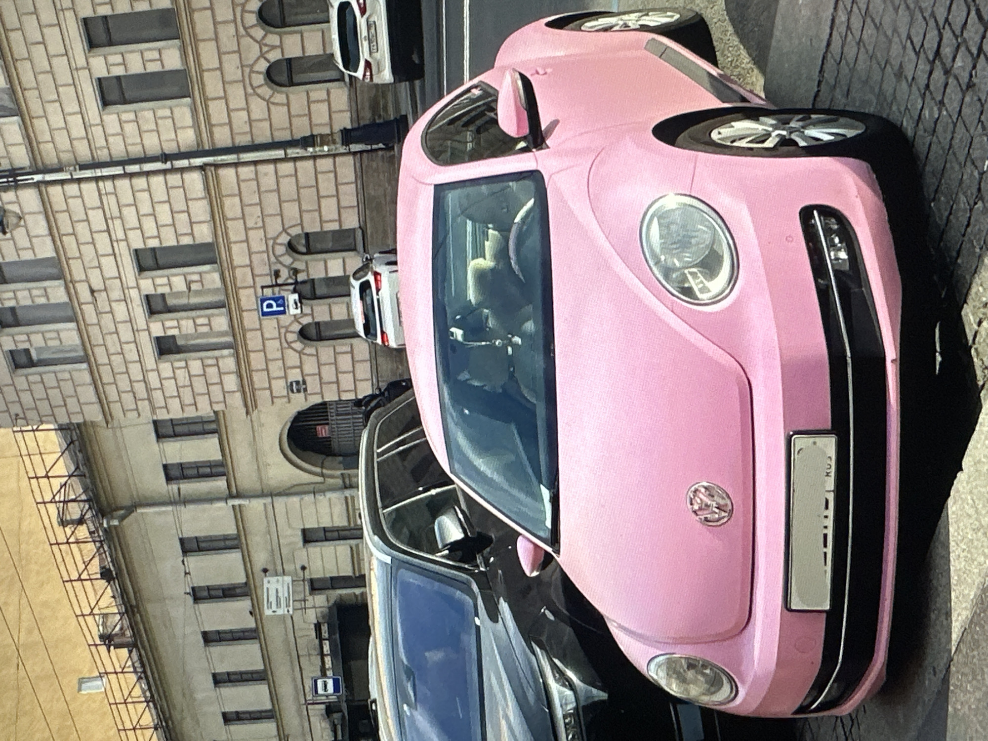 A pink Volkswagen Beetle is parked on a cobblestone street in front of a multi-story building with a light-colored facade. The Beetle has a black trim around its bumper and black side moldings. A black SUV is parked to the left of the Beetle. In the background, white cars are visible on the street, and there are signs indicating parking. The building in the background has arched windows on the lower level and rectangular windows on the upper levels, with some scaffolding on the roof. The scene appears to be in a European city, possibly Tallinn, Estonia, given the architecture and street environment.