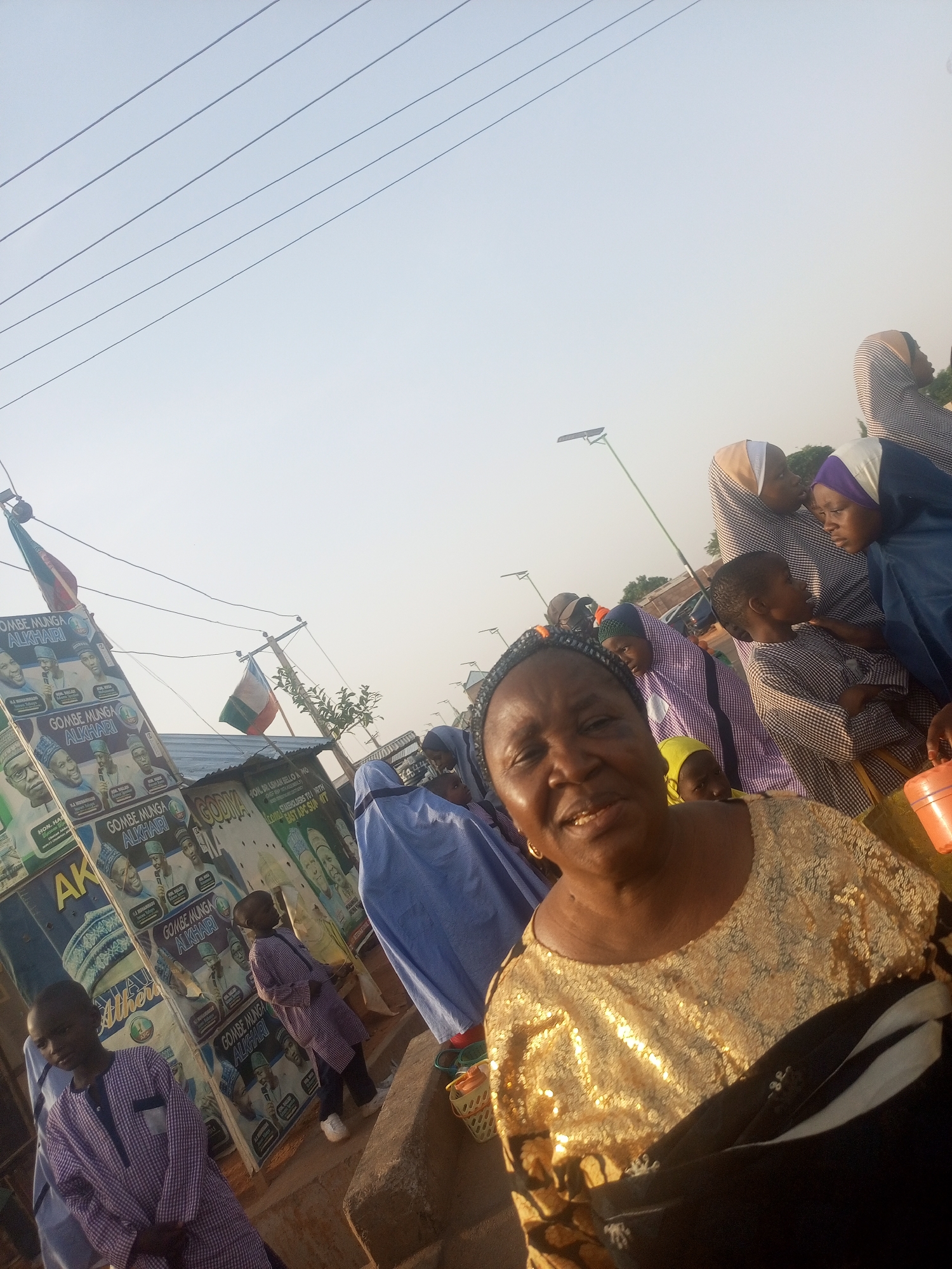 Here is a forensic description of the image:
**Scene:** An outdoor setting, possibly a street or public area in an urban or semi-urban location, featuring a group of people and political campaign materials.
**Foreground:** A middle-aged woman with a black head covering, wearing a shiny gold top with black accents. She is looking toward the camera with a slight smile.
**People:** Multiple individuals are present, including children. Some are wearing traditional or religious attire, such as hijabs in various colors and patterns. The clothing styles suggest a community with a mix of cultural influences.
**Objects:**
* Political posters displaying the faces of candidates, with the words "GOMBE MUNGA ALKHARI" prominently featured.
* A Nigerian flag.
* Power lines running across the sky.
* A street lamp.
* Other containers that appear to be plastic.
**Environment:** The background indicates a modest built environment with simple structures. The lighting suggests it could be either early morning or late afternoon.
**City/Location:** Information about the exact location and city of Unknown, Unknown, is not sufficient for a definitive identification. The environment looks like Nigeria, and the text indicates it is in Gombe State.