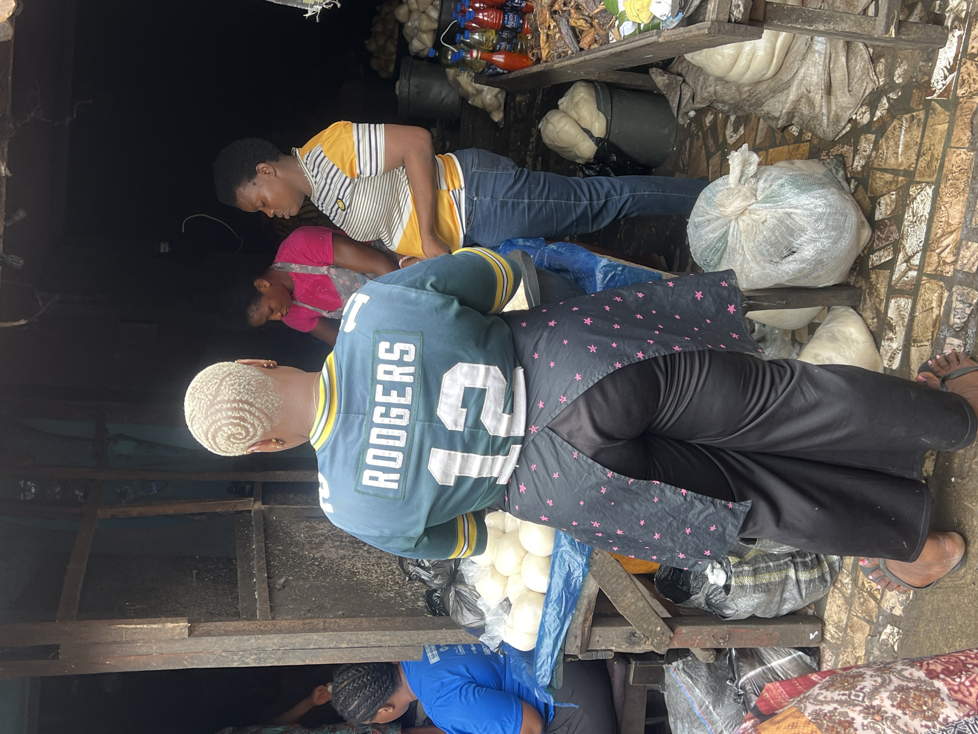 In Nkpor, Nigeria, a street market scene unfolds with several individuals engaged in commerce. The focal point is a person from the back, wearing a green and yellow football jersey with "RODGERS 12" emblazoned on it, paired with black pants and a black apron adorned with pink stars. This individual stands amidst a collection of goods. To the left, another person in a blue shirt is partially visible. In the background, two women are attending to a stall. One wears a pink shirt and a light-colored apron, while the other is in a striped yellow and white shirt with blue jeans, actively engaged with the items on display. The market environment is characterized by wooden structures, various sacks of produce, and a variety of bottled drinks and other goods arranged on shelves and the ground. The ground is paved with what appears to be brickwork, and it is wet, suggesting recent rain or cleaning. The overall impression is one of a bustling local marketplace.