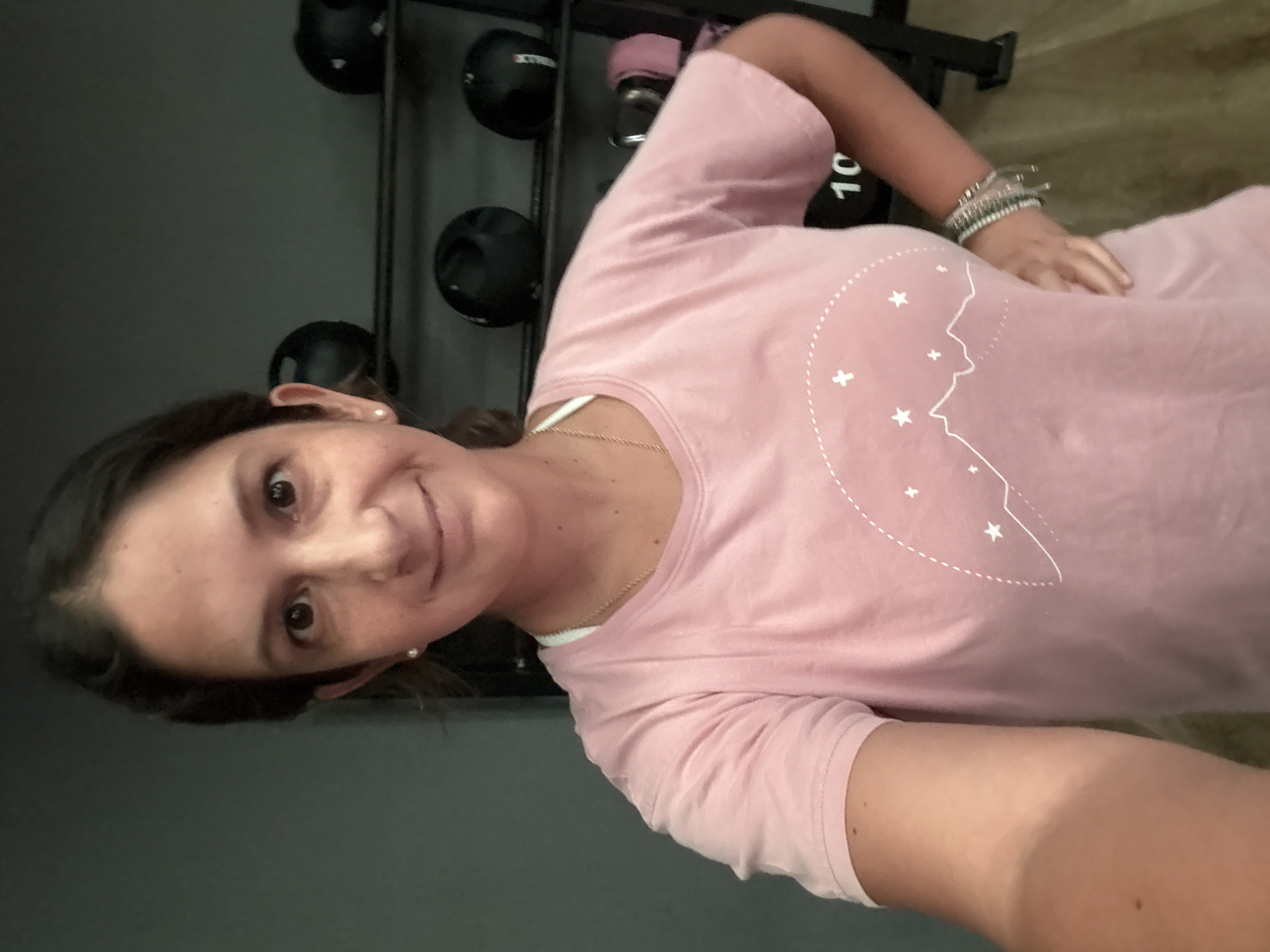 A woman with dark brown hair pulled back is shown from the chest up, taking a selfie in what appears to be a gym in San Isidro, Peru. She is wearing a light pink t-shirt with a white outline of mountains and stars. A delicate gold necklace and small pearl earrings are visible. Her left hand is on her hip, and she wears several bracelets on her right wrist. In the background, dark-colored exercise balls are stacked on shelves. The lighting suggests the photo was taken indoors during the day.