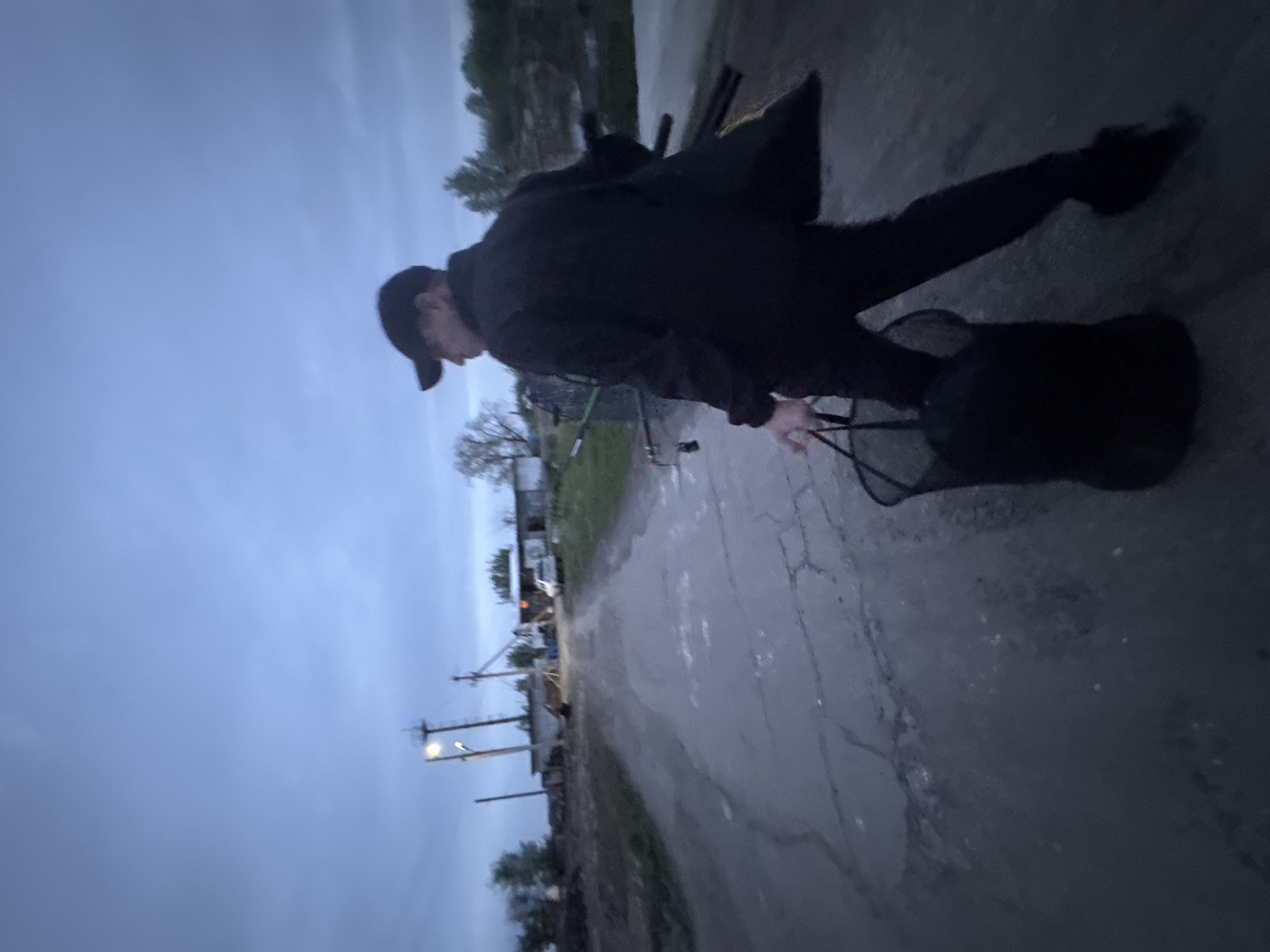 A person in dark clothing is seen walking along a paved road. The individual is wearing a cap and appears to be carrying a net or bag. The road has visible cracks and patches, suggesting wear or repair. In the background, there are utility poles with streetlights, trees, and buildings, indicating an urban or suburban setting. The sky is overcast, and the overall lighting suggests it may be dusk or dawn.