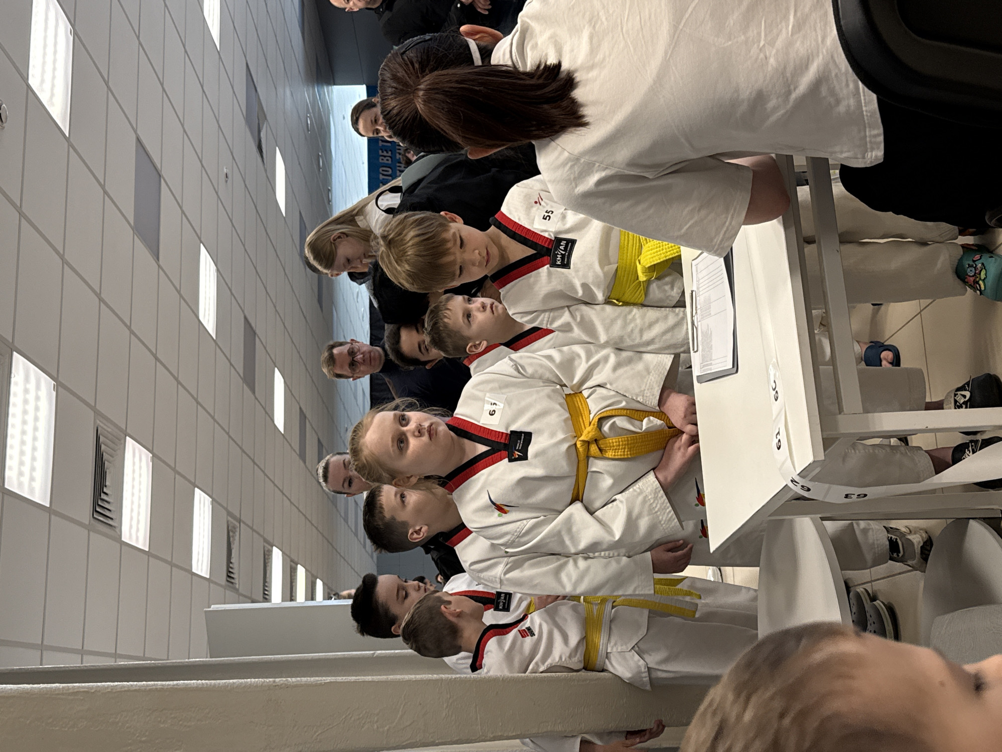 A group of people, mostly children, are lined up at a table. The children are wearing white taekwondo uniforms with yellow belts and identification numbers. Adults are standing behind them, some appearing to assist. The scene is indoors with a tiled wall in the background. No evidence suggests the location is Rostov-on-Don, Russia.