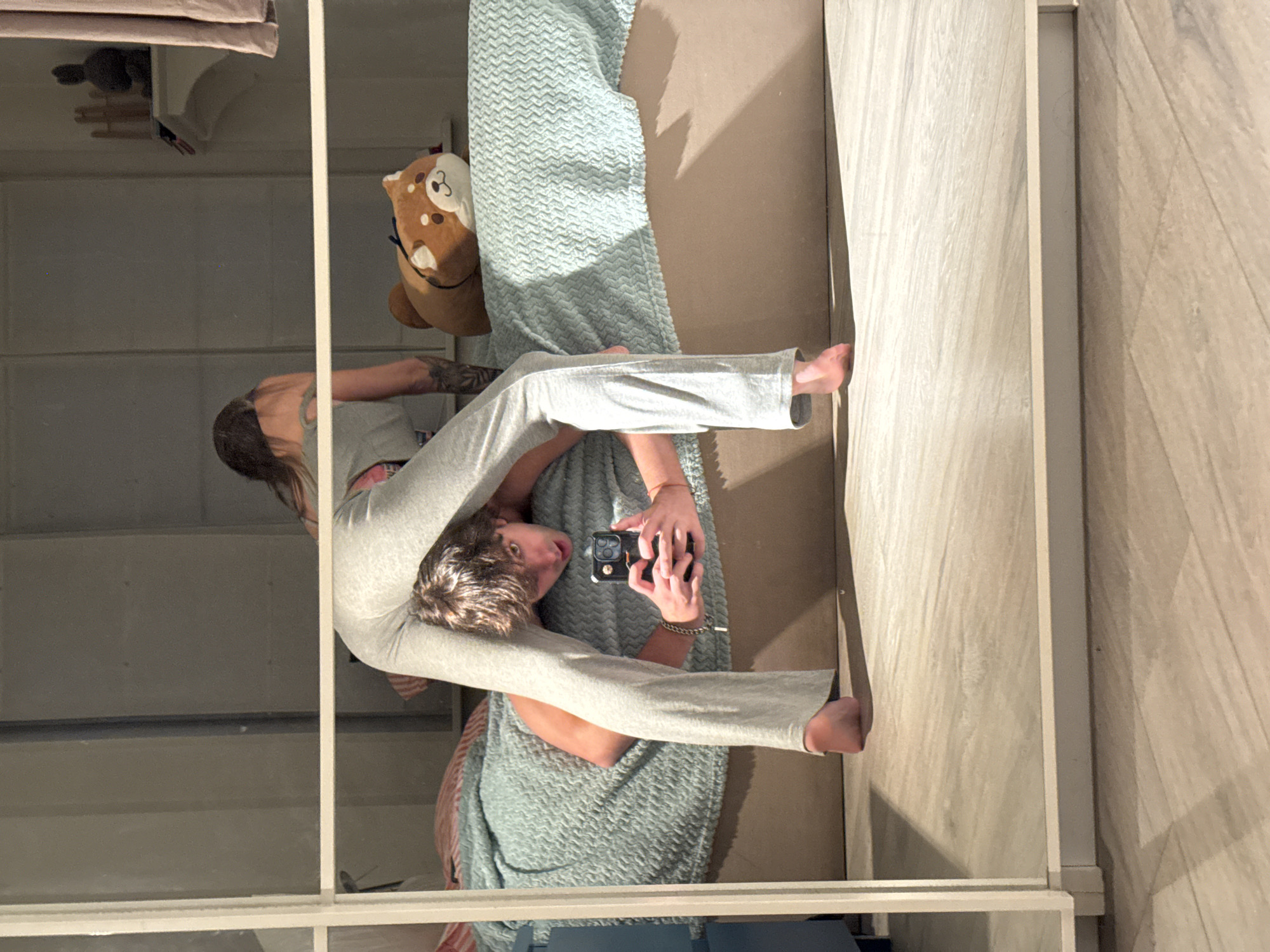 The image, taken in a bedroom in Moscow, Russia, shows a young man and woman in pajamas. The man is on his stomach on a bed, with his head bent backward as he holds a smartphone to take a selfie. The woman is in front of him, her body bent in a way that her legs are over his head. She is wearing gray pajamas, and he is also in gray pajamas. A stuffed dog toy is on the bed beside them. The room has wooden floors and a large mirror that reflects the scene.