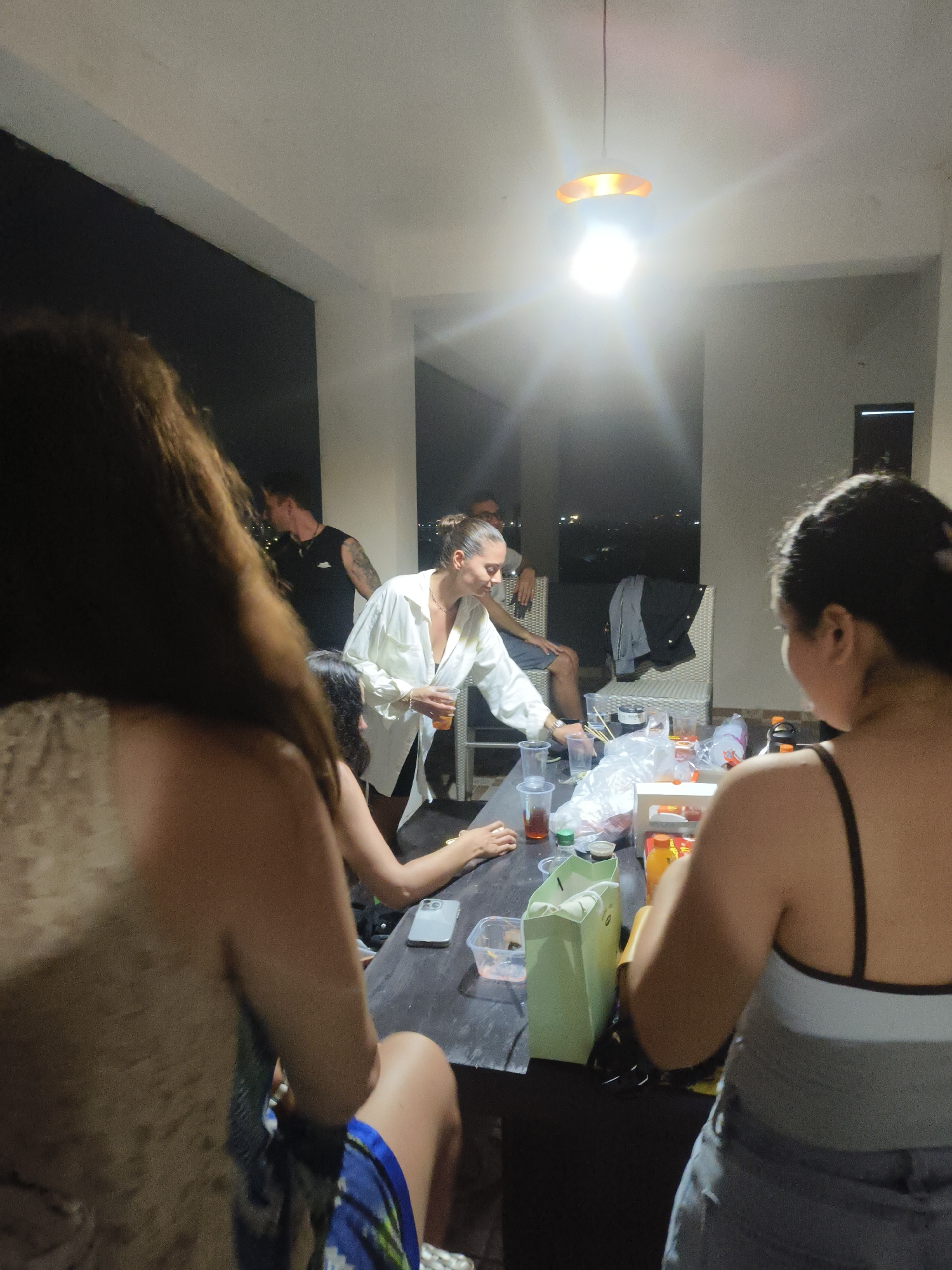 In Kuta Selatan, Indonesia, at night, several people are gathered around a dark-colored, rectangular table on an outdoor patio. A bright overhead light illuminates the scene. On the table are numerous clear plastic cups, some containing amber liquid, along with plastic bags, a light green paper bag, a yellow object, and what appears to be a phone. A woman in a white open-collared shirt and dark shorts is holding a cup and speaking. To her left, a person with dark hair is seated. In the foreground, a woman with long brown hair is partially visible, facing away from the camera. To the right, another woman with dark hair pulled back is facing the table. Further back, a man in a black tank top is interacting with the woman in the white shirt. Another person is seated in a white chair. The background shows a dimly lit urban or suburban landscape with distant lights.