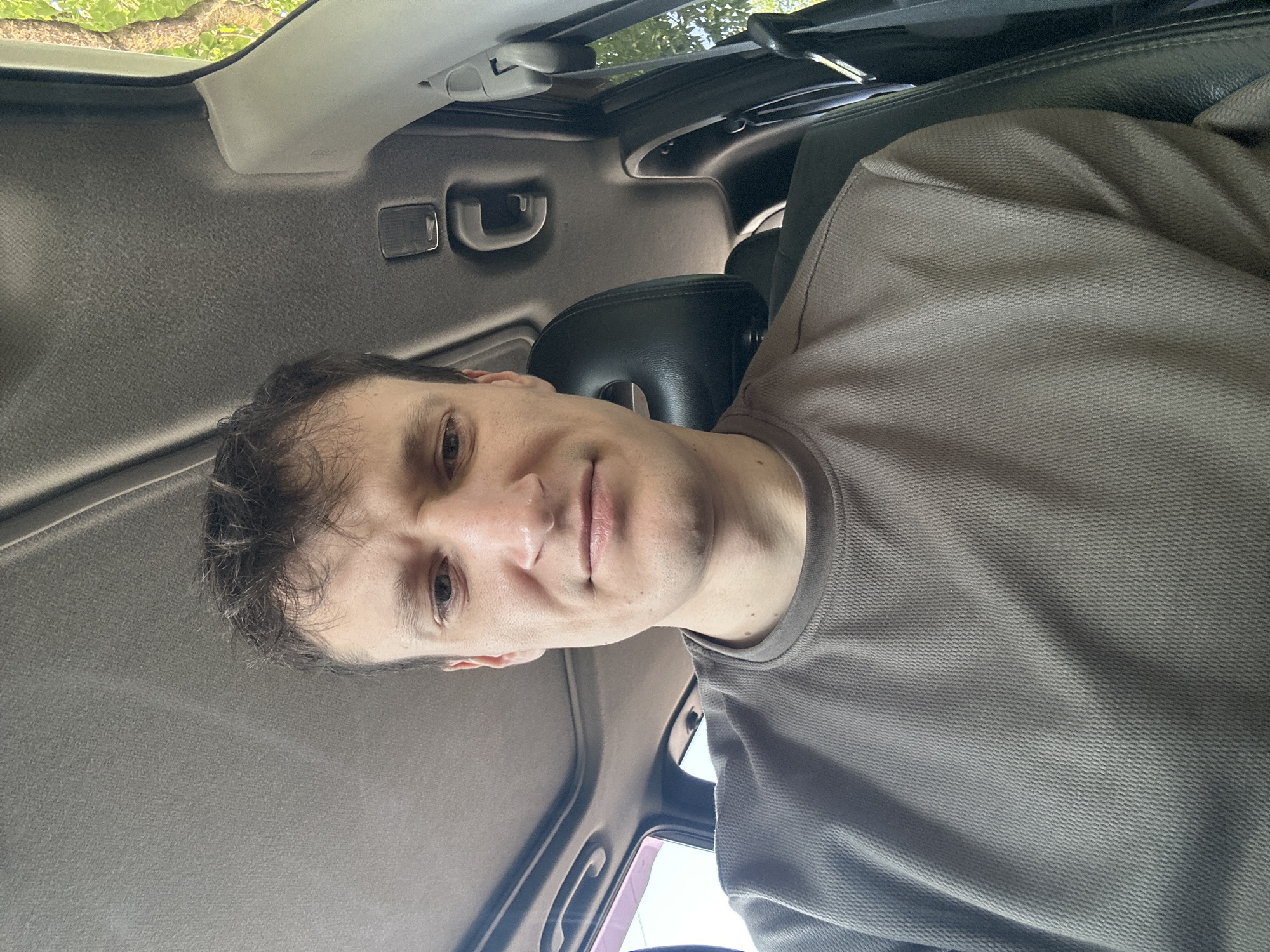 A man is sitting in the driver's seat of a car, looking at the camera. The car's sunroof is open, and sunlight is visible. The man is wearing a gray t-shirt. The interior of the car is visible, including the dashboard and the headrest of the passenger seat. The background shows trees and foliage, suggesting the car is parked outdoors, possibly in a residential area or on a street in Makiv, Ukraine. The man appears to have a neutral expression on his face.