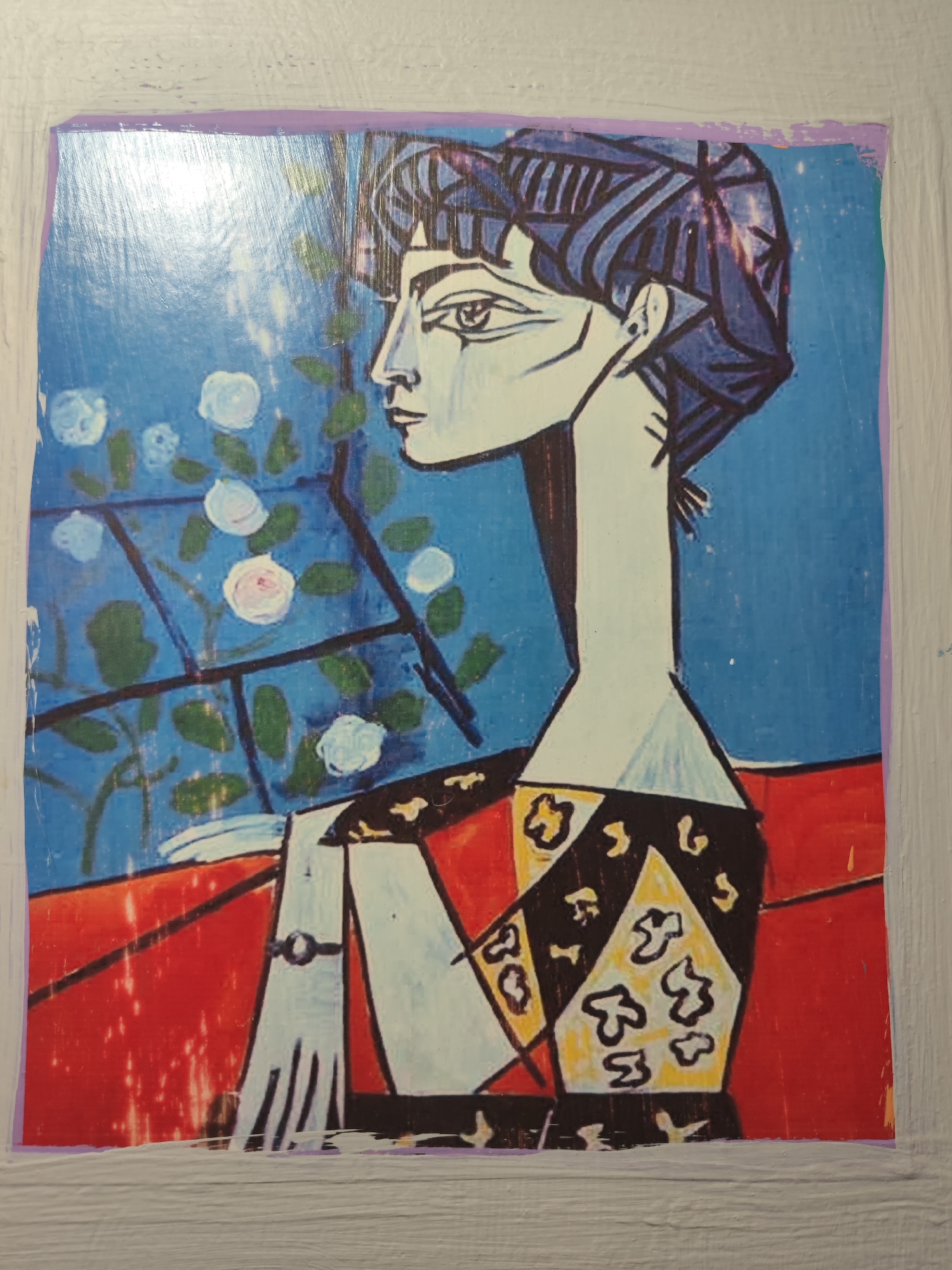 The image is a postcard of a painting in the cubist style, depicting a woman with dark hair and sharp, angular features. She is wearing a patterned dress with a red background. The painting is framed by a purple border and is placed on a light gray, textured surface, possibly a wall. The postcard itself is slightly creased and worn. The background of the painting shows a blue sky with white and pink roses against a window frame. The location is identified as Tuzdybastau, Kazakhstan.