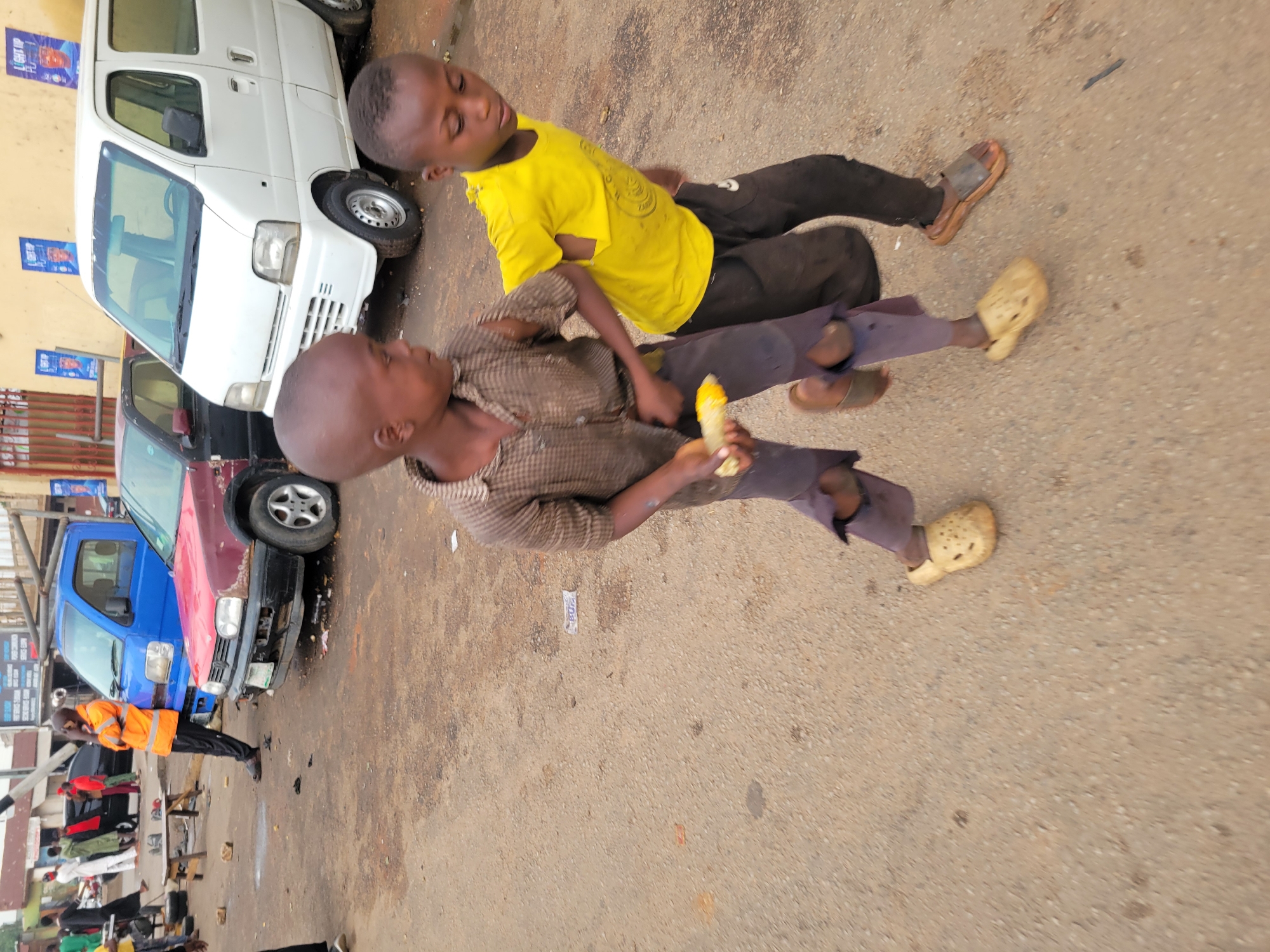 Two African children are sitting on the ground next to a busy street scene in Jos, Nigeria. One child wears a yellow shirt and black pants, while the other wears a brown shirt and purple pants. The child in brown pants is holding what appears to be a piece of corn. Both children are wearing sandals. Nearby are several parked vehicles including a white van, a red truck, and a blue truck. Pedestrians are walking in the background.