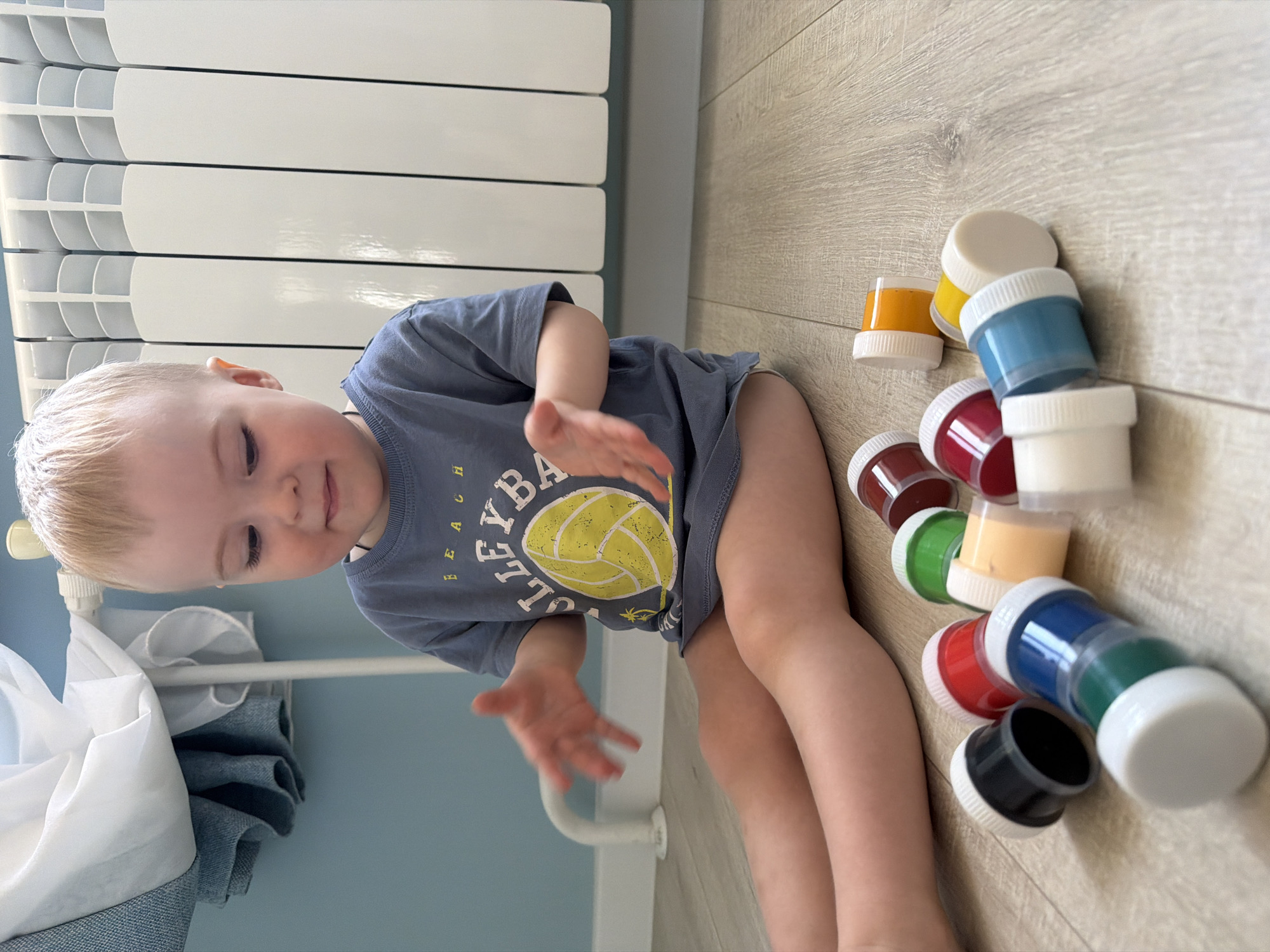 A young child with blonde hair, wearing a grey t-shirt with a volleyball design, sits on a light wood-grain floor next to a collection of small, stacked paint containers. The containers are various colors, including yellow, blue, red, green, and black, all with white lids. Behind the child is a white radiator and, to the left, a blue wall with white curtains. While the location is unspecified, the description is consistent with a possible indoor residential setting. There is no information to suggest that the location is Votkinsk, Russia.