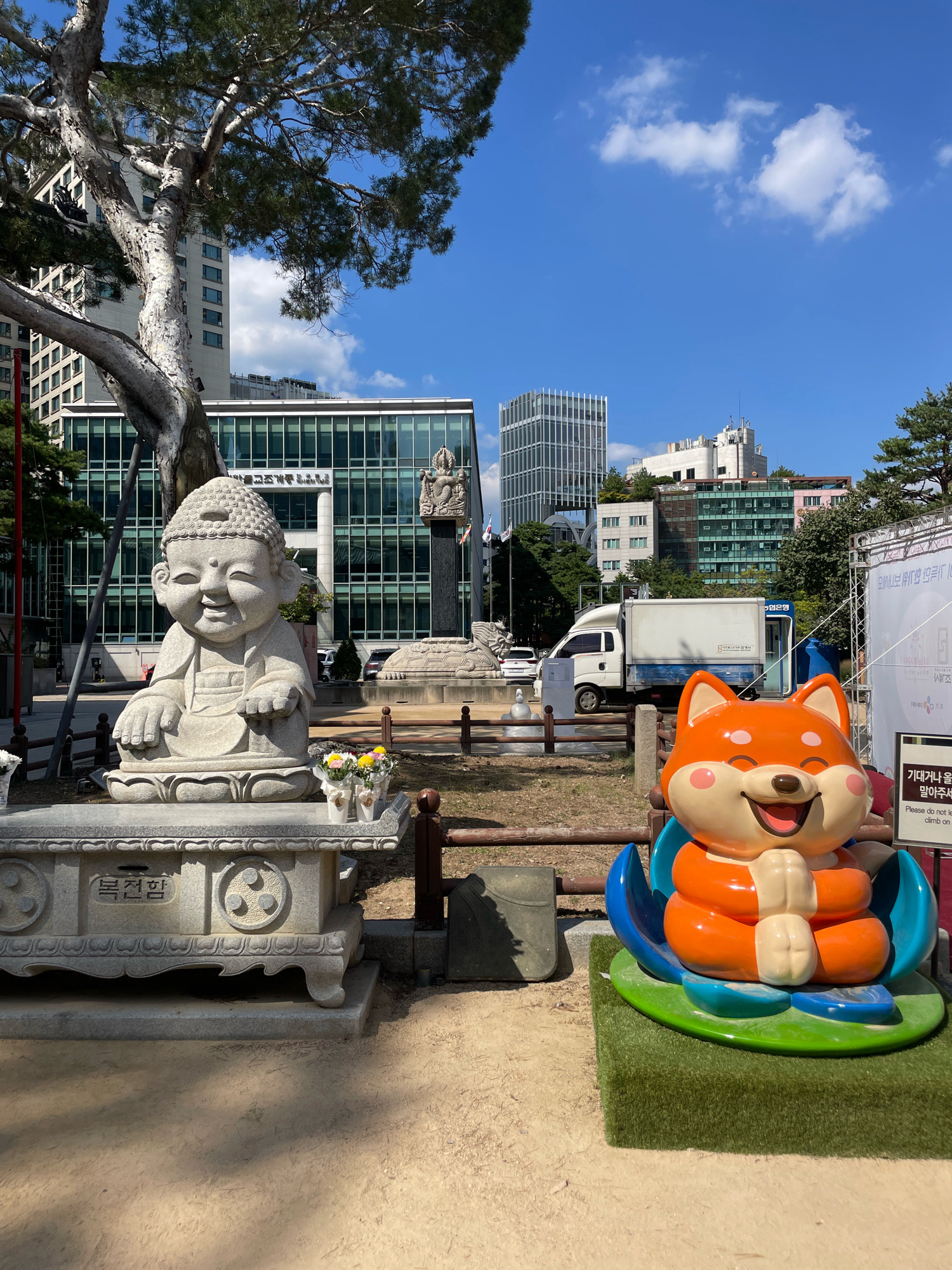 This is a photograph taken outdoors in Seoul, South Korea. The scene features a stone statue of a smiling, seated Buddha in the foreground on the left. To the right of the Buddha statue, there is a large, brightly colored orange cartoon fox statue sitting in a blue and green lotus flower. Both statues are placed on a sandy area with a wooden fence in front of them. Behind the statues, there is a modern building with a glass facade and a tall tree with a gnarled trunk. Further in the background, several other buildings of varying heights and architectural styles are visible, along with a clear blue sky with some white clouds. A white truck with a blue stripe is parked behind the statues. A sign with Korean text and an English phrase "Please do not lean on or climb on it" is visible near the fox statue. The overall impression is a blend of cultural elements and modern urban landscape.