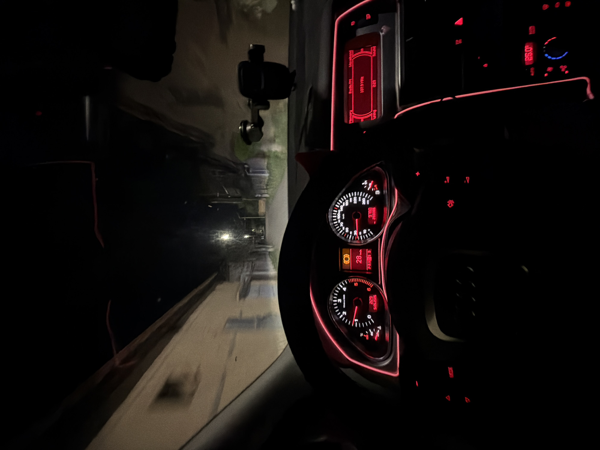 The image depicts the interior of a car at night, specifically the dashboard and part of the road visible through the windshield. The dashboard features a speedometer and tachometer with red illumination, along with a radio display and various controls. A phone holder is mounted on the windshield. The road outside is lit by streetlights, suggesting a residential or urban area. The radio display shows "Radio FM 107.9 MHR". A yellow light symbol is present that could indicate "Low Tire Pressure". The odometer reading is 5.05.2026 km, suggesting a reference to a future date. The gear selector displays "PRNAS 3". There is no verifiable indication that the image was taken in Byala Cherkva, Bulgaria.