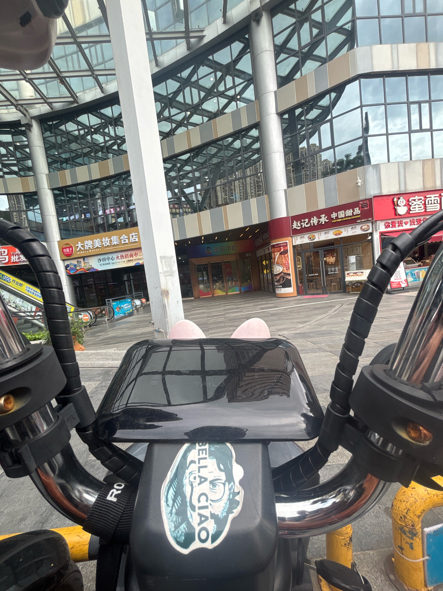 The image shows a close-up view of the handlebars of an electric scooter parked outdoors in front of a modern commercial building. The building features a glass facade with a geometric grid pattern and reflects the sky and surrounding structures. Multiple storefronts are visible at the ground level, some with red and white signage, indicating commercial establishments. One sign reads "Zhao Ji Cheng Pin Zhong Guo Tian Pin" with Chinese characters and the English translation "Zhao Ji Inherited China Sweet Food". Another sign, partially visible, appears to advertise a supermarket or retail store. The ground is paved with square tiles. The scooter's handlebars are black with a coiled cable wrap. A sticker depicting a face and the text "BELLA CIAO" is adhered to the scooter's console. Two small pink, ear-shaped decorations are attached to the top of the console. Yellow and blue metal poles are visible in the foreground, likely part of street furniture or barriers. The scene appears to be in Nanhai District, China, based on the context provided.