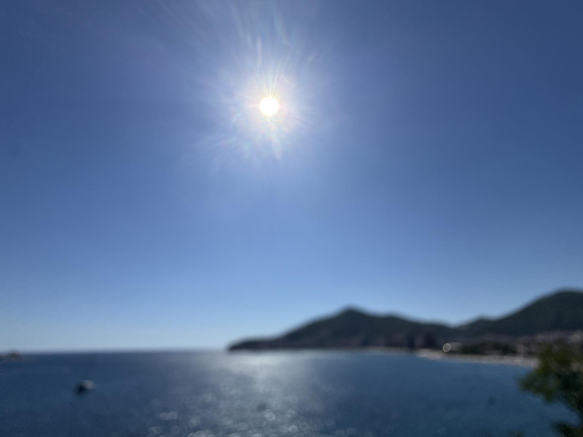 The image captures a bright, sunny day over a coastal scene. The sun is high in a clear, vivid blue sky, casting a strong glare. In the middle ground, the deep blue sea stretches towards a hazy, dark silhouette of a mountainous coastline, likely representing the city of Budva, Montenegro. Buildings are faintly visible along the shore, nestled against the hills. The water's surface is disturbed by sunlight, creating shimmering, blurred reflections. A small, indistinct boat is visible on the left side of the sea. The overall impression is one of a serene, sun-drenched coastal landscape, with the city of Budva forming a backdrop.