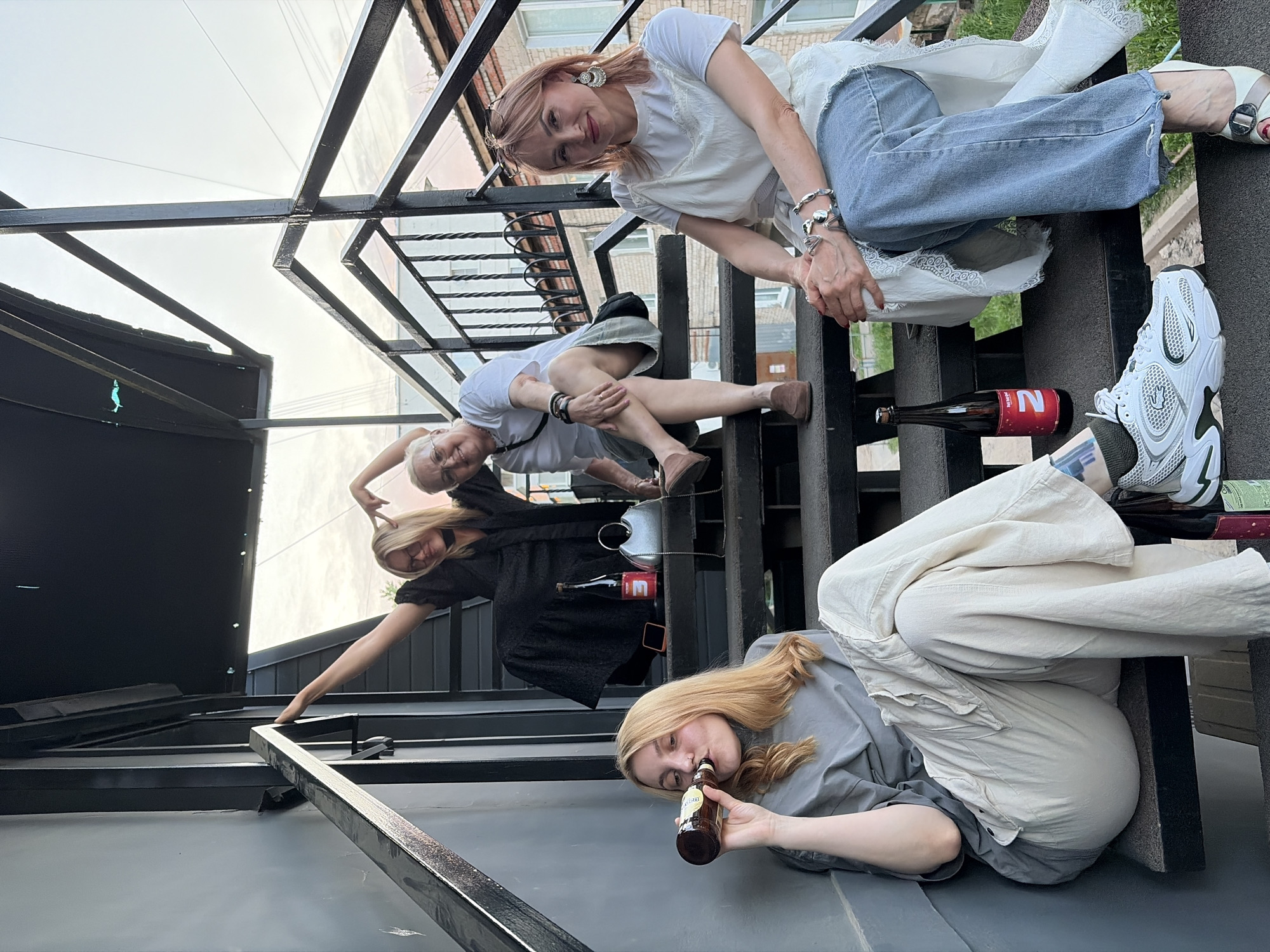 Four women are sitting and lying on a black metal staircase. One woman in the top right of the frame is wearing a white shirt and blue jeans. Below her is a woman with short gray hair wearing a white t-shirt and gray shorts. The next woman down is wearing a black shirt. The woman at the bottom of the frame is wearing a gray shirt and beige pants, and drinking from a brown bottle. There are bottles of beer on the stairs as well. The photo was taken in the city of Chaikovsky, Russia.