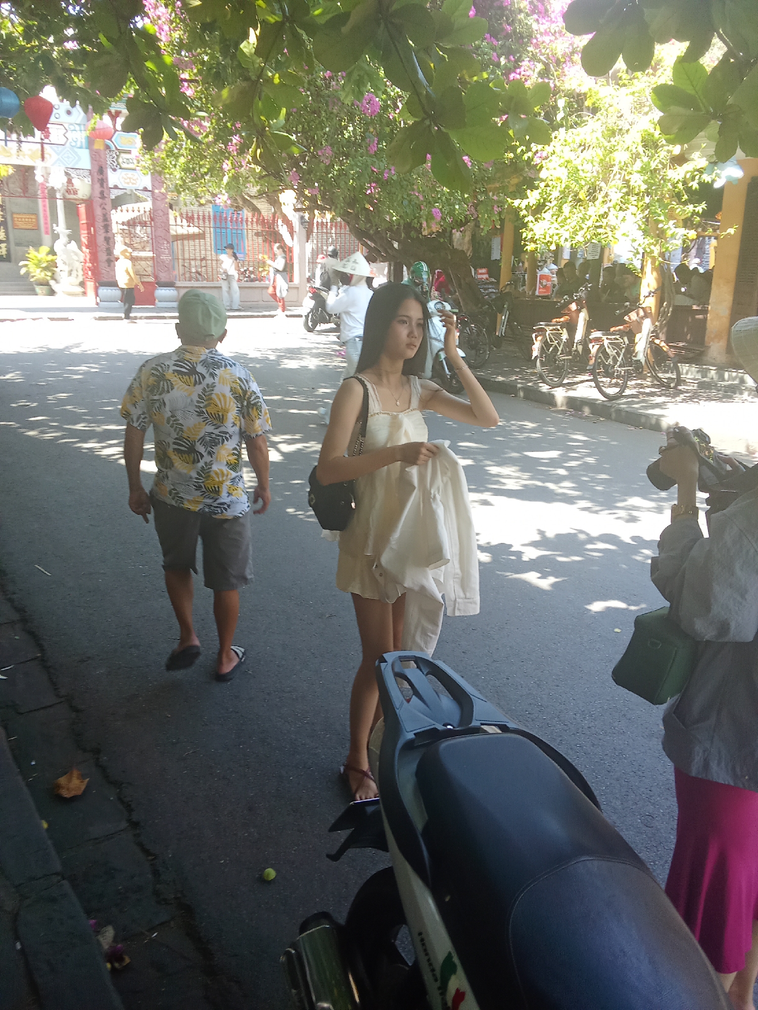 The image depicts a street scene in Hội An Ward, Vietnam, likely during the daytime given the sunlight and shadows. In the foreground, a person wearing a colorful patterned shirt and shorts, with a green cap, walks away from the camera. To their right, a young woman in a light-colored dress is holding a white garment and looking towards the left. To the far right, another individual is visible, wearing a grey jacket and a bright pink skirt, holding a camera. The background shows a street lined with buildings and trees, with bicycles and motorcycles parked along the roadside. The architecture suggests a historic urban setting. The overall atmosphere is casual and appears to be a public area.