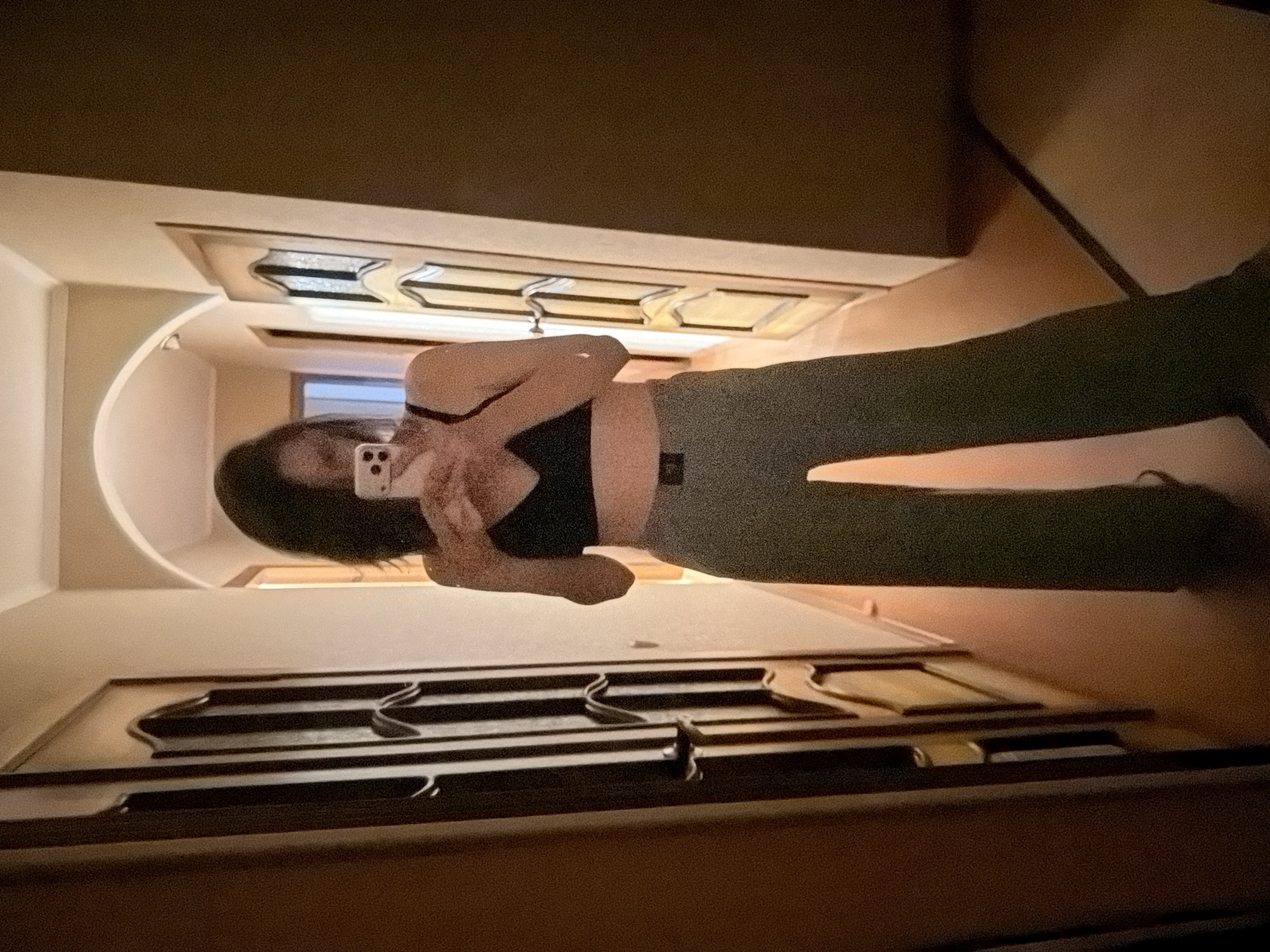 A person is taking a mirror selfie indoors. The person is wearing a black top and green pants. The setting appears to be a bathroom. The city and precise location are Unknown.