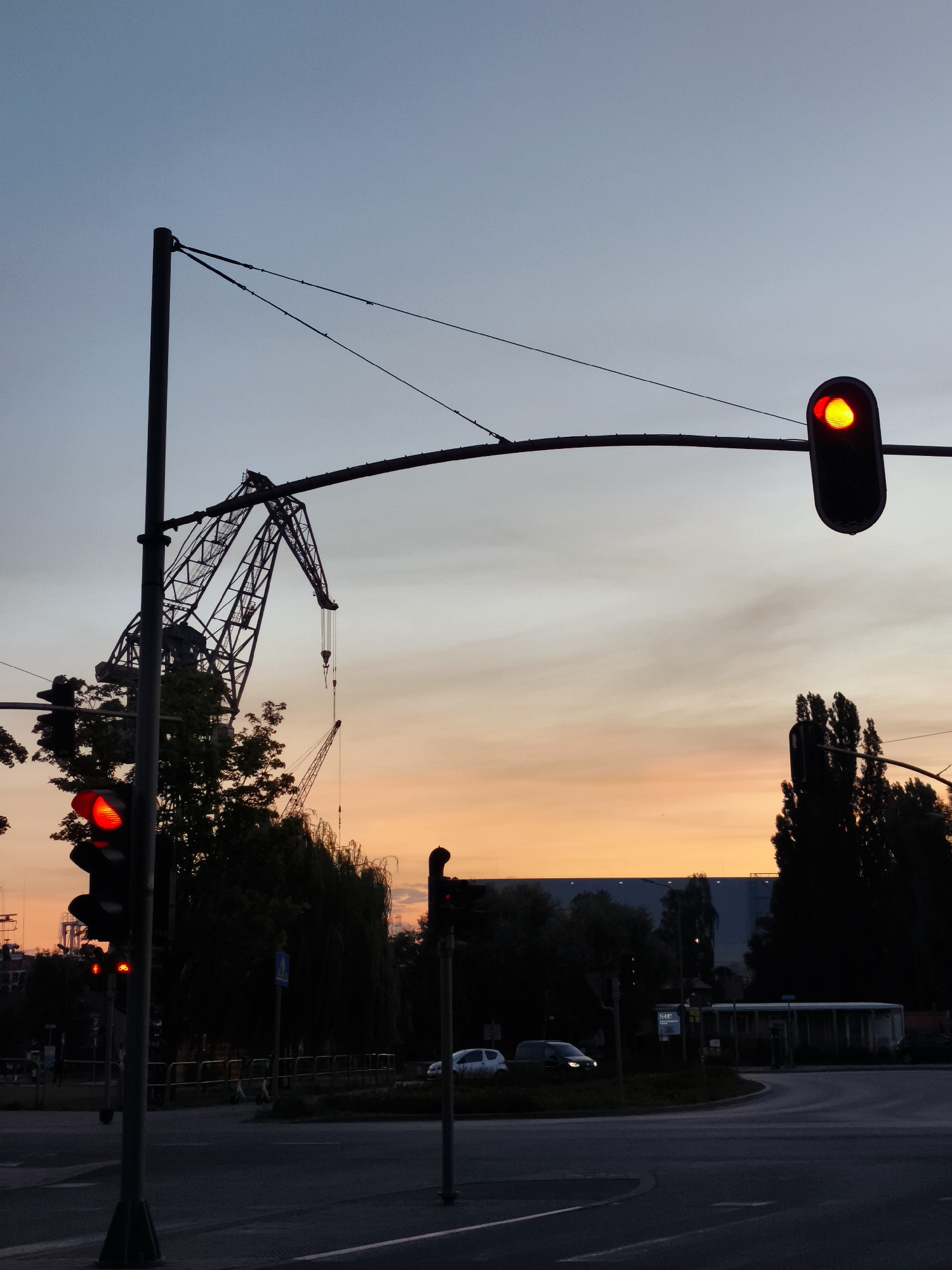 The image depicts a dusky urban scene with a vibrant orange and blue gradient sky. In the foreground, a silhouetted traffic light displays a red signal. To the left, another traffic light is also illuminated red, with a smaller red signal below it, possibly indicating a pedestrian crossing or a separate lane. A large, skeletal industrial crane, also in silhouette, dominates the mid-ground, with a smaller crane visible behind it. Trees and other structures are obscured by darkness. The scene suggests a port or industrial area, consistent with the city of Gdańsk, Poland, known for its maritime and industrial heritage. The road surface is visible in the foreground, with faint lane markings. Two cars are partially visible on the road. The overall impression is one of twilight settling over an active industrial landscape.
