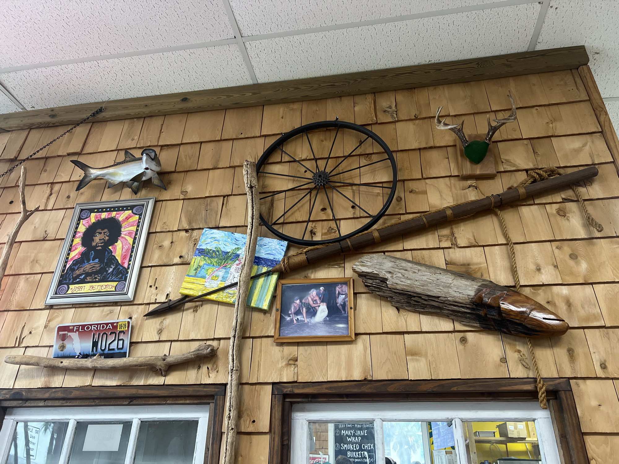 This image displays a wall of a restaurant in Jacksonville Beach, Florida, United States of America, adorned with various nautical and rustic decor. Mounted on the wooden-shingled wall are a taxidermied fish, a framed picture of Jimi Hendrix, a Florida license plate, a wagon wheel, deer antlers, a spear, a painting of a beach scene, a framed photograph of people pulling a net from the water, and a wooden fish sculpture. A rope hangs down to the right of the wooden fish sculpture. Two windows with wooden frames are visible at the bottom of the image, showing a reflection of the interior of the restaurant, including a menu.
