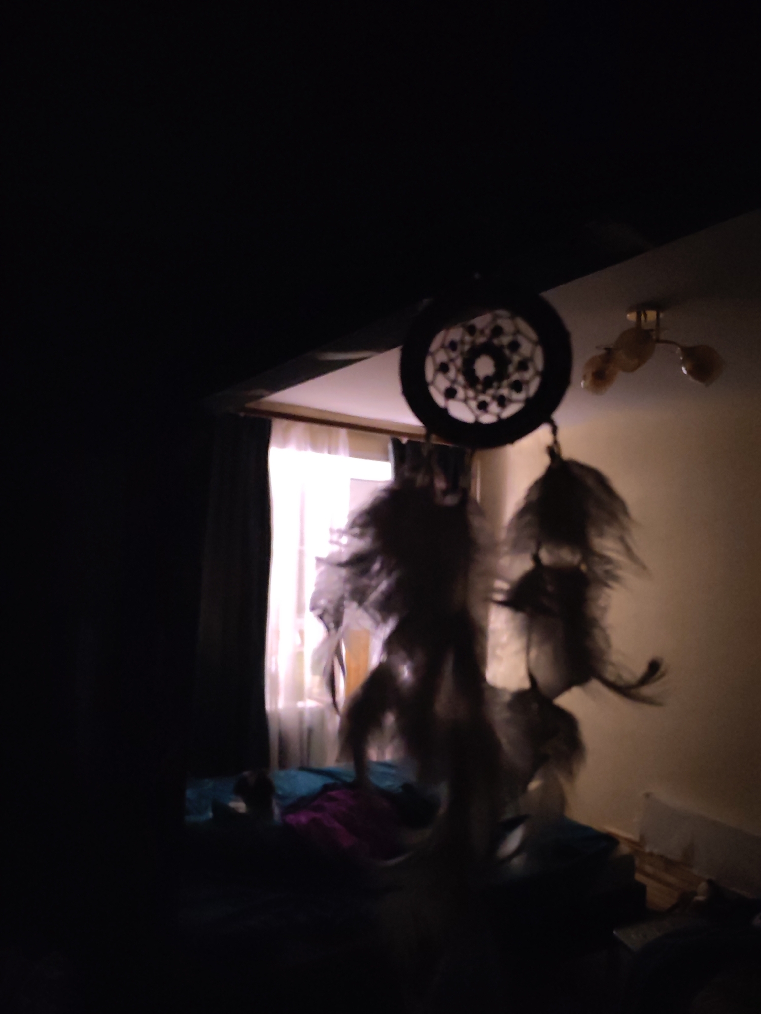 The image shows a dimly lit room, likely a bedroom, in Izhevsk, Russia. A dreamcatcher hangs in the foreground, positioned between the viewer and a window covered with sheer curtains. The bed is visible in the middle ground, draped with blue and purple covers. In the background, a wall is visible, illuminated by a ceiling light fixture with multiple globe-shaped lamps. The overall atmosphere is dark, obscuring details.