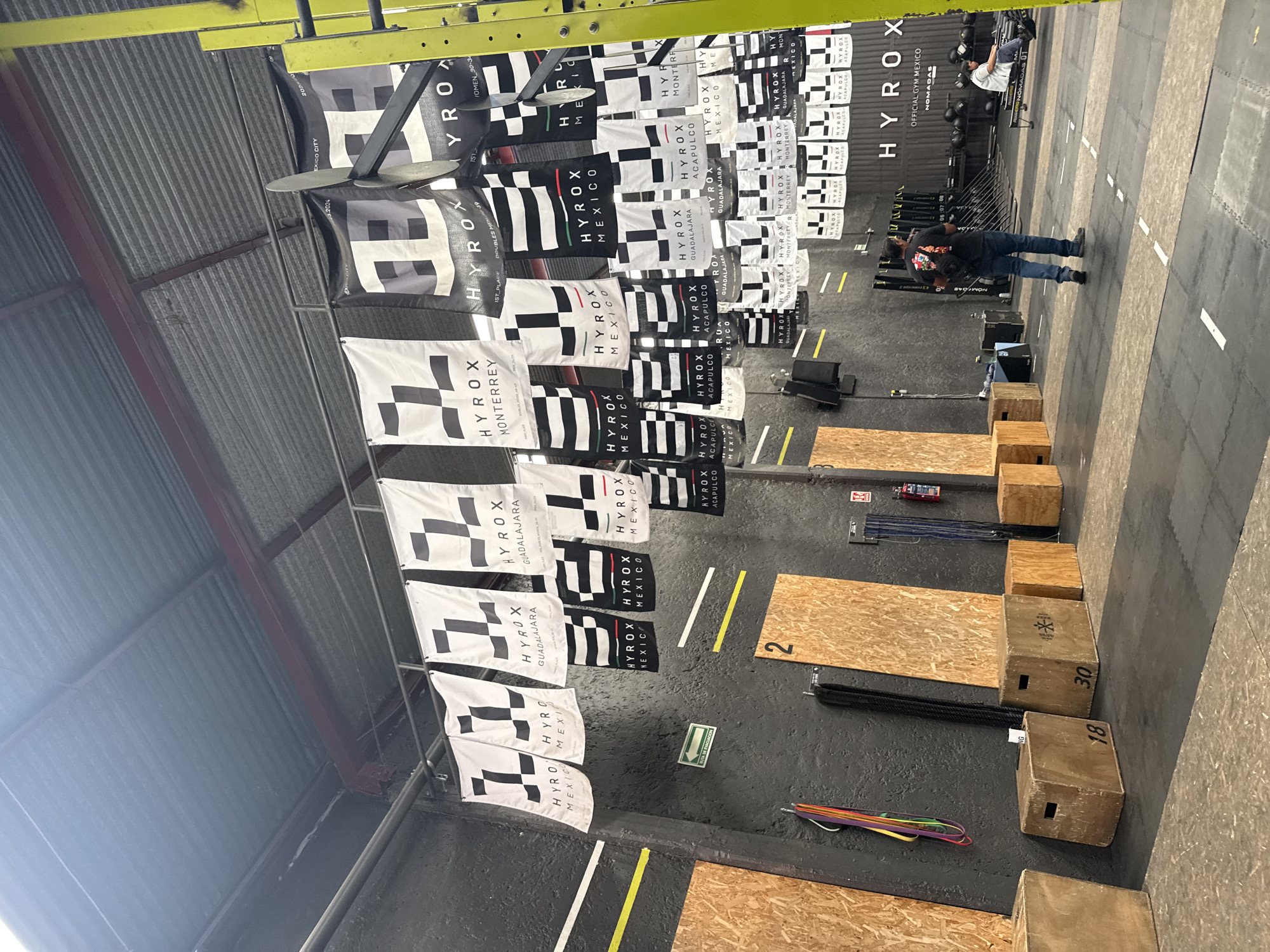The image displays the interior of a fitness facility or gym in Mexico City, likely set up for a HYROX event or competition. The setting features a corrugated metal ceiling with yellow support beams and a dark floor marked with white and yellow lines.
Dominating the left side are numerous black and white banners, many featuring a checkered pattern and the prominent "HYROX" logo. These banners bear text indicating various locations and categories, such as "MEXICO CITY," "MONTERREY," "GUADALAJARA," "ACAPULCO," and general "MEXICO." Specific competition details like "WOMEN 30-34," "DOUBLES MEN," "1ST PLACE," and a date "3.2024" are also visible. A ceiling fan is suspended among the banners.
The floor is equipped with fitness gear, including several wooden plyometric boxes, some marked "18" and "30," thick ropes, and colorful resistance bands. In the background, exercise machines resembling Concept2 rowers or ski ergs are visible, along with a rack of black weights. Two individuals are present: one person wearing a black t-shirt and blue jeans stands in the foreground, while another is further back, appearing to be on a piece of equipment. A wall sign reads "OFFICIAL GYM MEXICO NOMADAS," and an exit sign with a left-pointing arrow is also present. The scene strongly suggests a fitness competition or a training facility for such events.