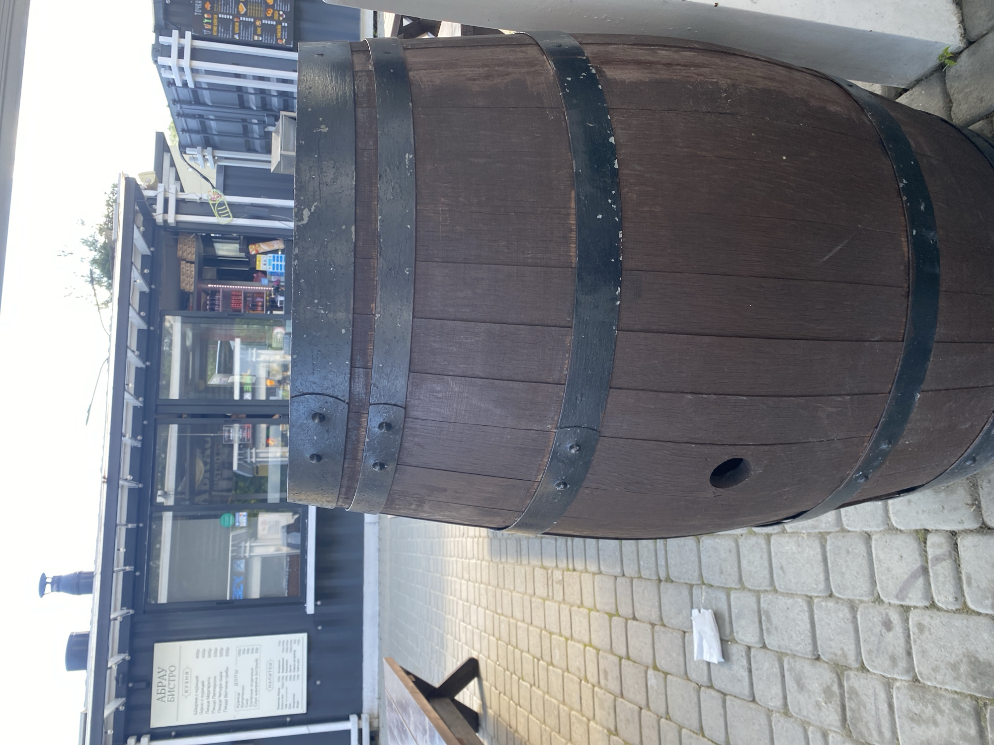 This image depicts an outdoor scene, likely in a commercial or promenade area in Abrau-Dyurso, Russia, during clear daylight hours.
The main subject is a large, dark wooden barrel positioned horizontally in the foreground and extending across the right side of the frame. It is adorned with several dark metal bands, secured by visible rivets, and features a small, circular opening on its side. The barrel rests on light-colored, rectangular paving stones that cover the ground. A small piece of white litter is visible on these stones near the barrel.
In the mid-ground, to the left of the barrel, is a modern-looking food stall or bistro. This structure is predominantly black with white decorative elements and large glass windows that reflect the surroundings. A prominent menu board is affixed to the bistro's exterior. Visible text on the board includes "АБРАУ БИСТРО" (Abrau Bistro) and "КУХНЯ" (Cuisine). The menu lists items such as "Шаурма с курицей" (Chicken Shawarma), "Парос с курицей" (Chicken Gyro/Paros), and several pizzas including "Пицца Маргарита" (Pizza Margherita), "Пицца Пепперони" (Pizza Pepperoni), "Пицца Четыре сыра" (Four Cheese Pizza), and "Пицца Ветчина-грибы" (Ham and Mushroom Pizza), alongside prices in Rubles. A section for "НАПИТКИ" (Drinks) is also present. Another sign overhead reads "ТОЧКА" (Point/Spot).
A wooden bench is partially visible in the lower-left portion of the image. The bright sky and visible shadows indicate it is daytime with strong sunlight. No people are present, suggesting a quiet moment.