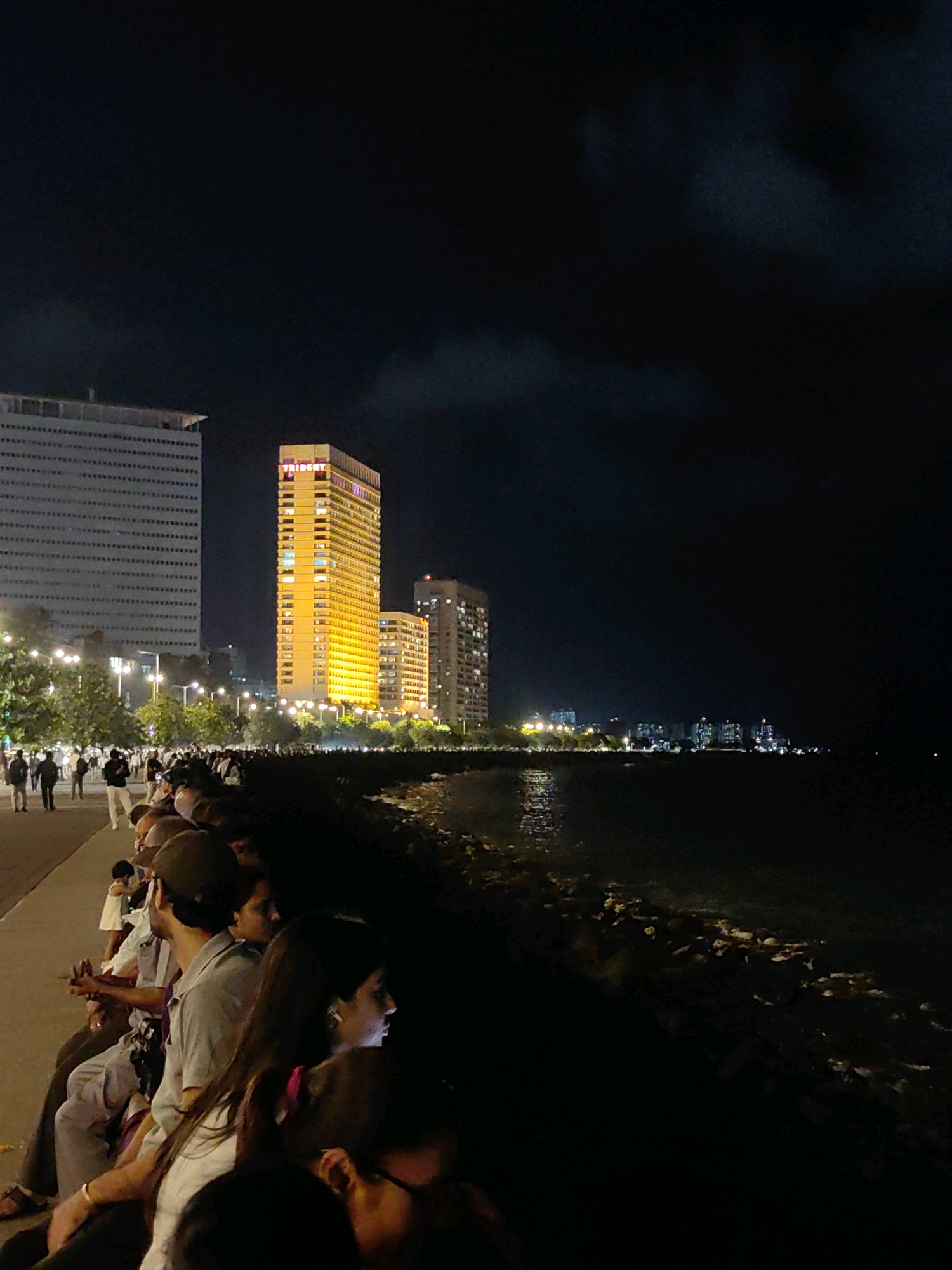 The image depicts a nighttime scene at Marine Drive, Mumbai, India. A long line of people is seated along a seawall, facing away from the camera and towards the ocean. Beyond the people, a well-lit promenade with streetlights and trees stretches into the distance, leading to several prominent buildings. The most striking building is a tall, slender skyscraper illuminated in a bright golden-yellow hue, with the word "TRIDENT" visible near its top. To its left is a wider, less brightly lit, multi-story building with a grid-like pattern of windows. Further in the distance, more buildings with illuminated windows form a city skyline against the dark night sky. The ocean is visible to the right of the frame, with subtle reflections of the city lights on its surface. The overall atmosphere is one of a popular urban gathering spot at night.